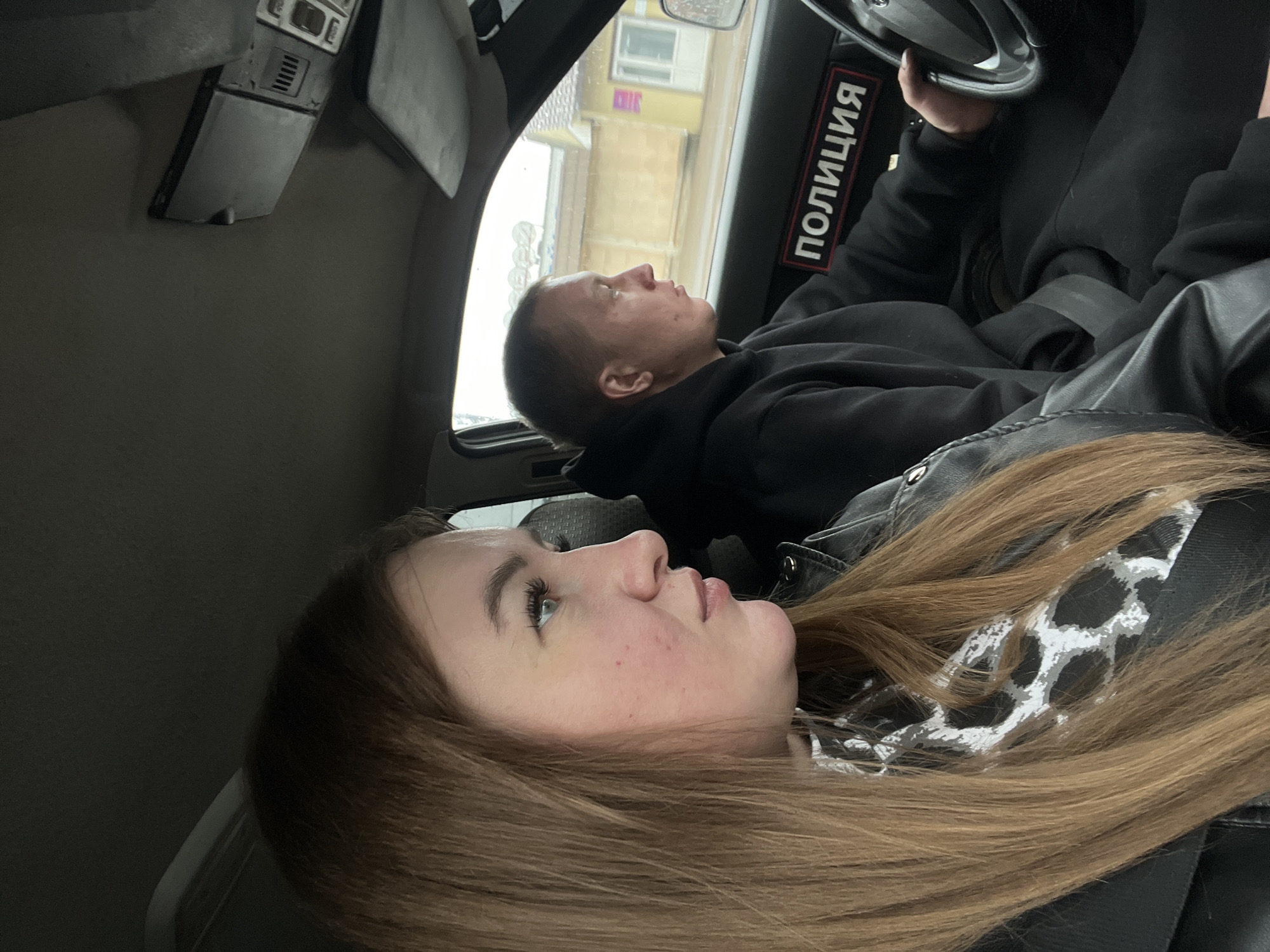 Two people are in a car. One person is driving and the other is a passenger. The driver appears to be a male with short hair and is wearing a black sweatshirt. The passenger appears to be a female with long brown hair and is wearing a black jacket with a white and grey patterned scarf. The word "ПОЛИЦИЯ" (Police) is seen on the door. The scene is in an urban environment, potentially in the city of Chaikovsky, Russia.