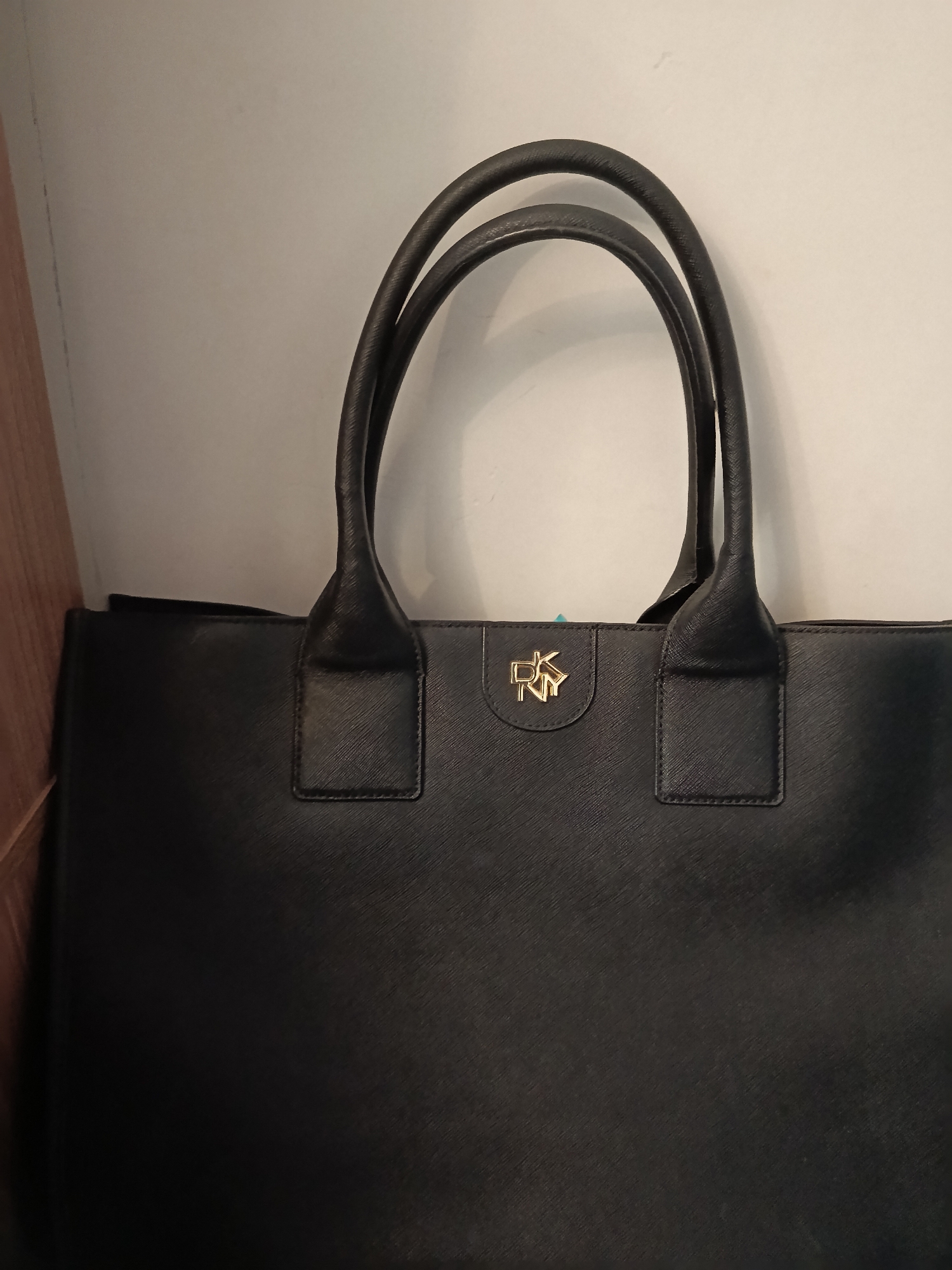 A black leather DKNY tote bag is centered in the frame, suspended by its two handles. The bag has a textured surface. A gold DKNY logo is visible on the upper front of the bag, positioned between the two handle attachments. The background is a plain, light-colored wall and a portion of a wooden surface is visible on the left edge of the frame.