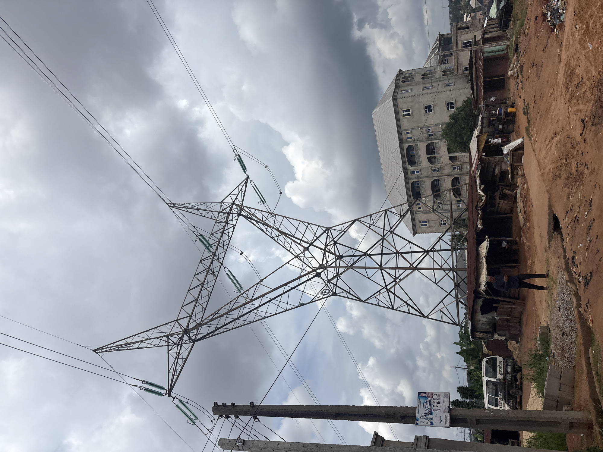 The image depicts an outdoor scene in Nkpor, Nigeria, dominated by a large electricity pylon and transmission lines against a cloudy sky. In the background, a multi-story building with numerous windows stands alongside informal structures, likely shops or homes, constructed from corrugated metal and wood. A dirt road runs along the right side of the frame, with piles of debris and some vegetation visible.
A person is standing near the informal structures, looking towards the left. A white van is parked nearby. The sky is overcast with dramatic grey clouds, suggesting a potentially stormy or gloomy day. The overall atmosphere is one of an urban or semi-urban environment with a mix of modern and informal infrastructure.
On a pole in the foreground, a poster is visible with text that appears to be an advertisement. While difficult to read clearly, some words like "CHALLENGE" and "EDUCATION" might be discernible, along with multiple faces of people. The time of day is indeterminate due to the cloud cover, but the lighting suggests it is daytime. The location cues of the informal structures, the type of van, and the overall urban landscape point to a Nigerian setting.