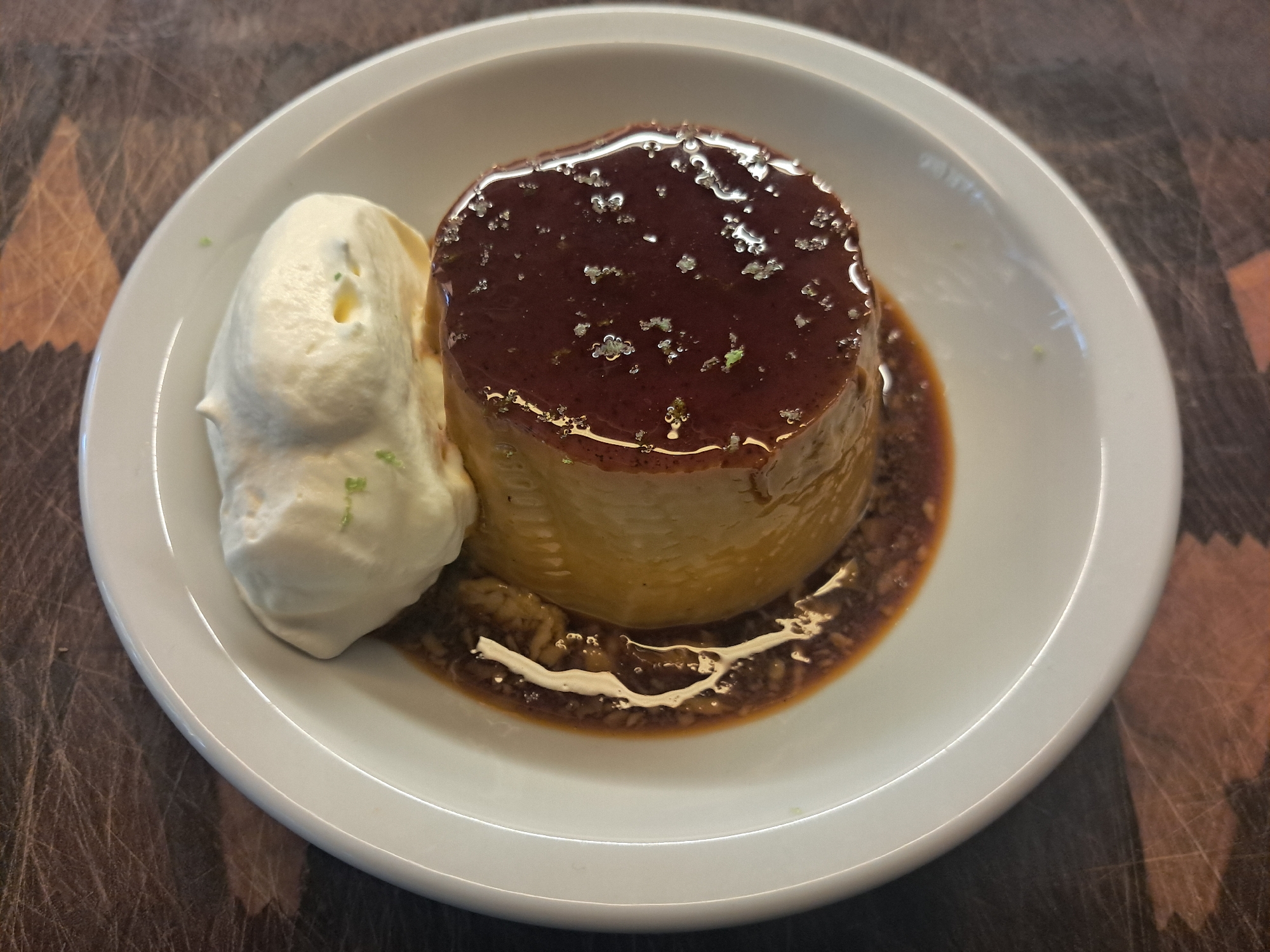 The image shows a dessert, likely a flan or crème caramel, presented on a white plate. The flan is a cylindrical shape and sits in a pool of caramel sauce. It is topped with what appears to be sugar crystals or possibly edible silver flakes. To the left of the flan is a scoop of whipped cream, garnished with what might be lime zest. The plate rests on a wooden surface with a distinctive zigzag pattern. The image does not contain information or depict anything that would allow inferring the location in the city of Vicente López, Argentina.