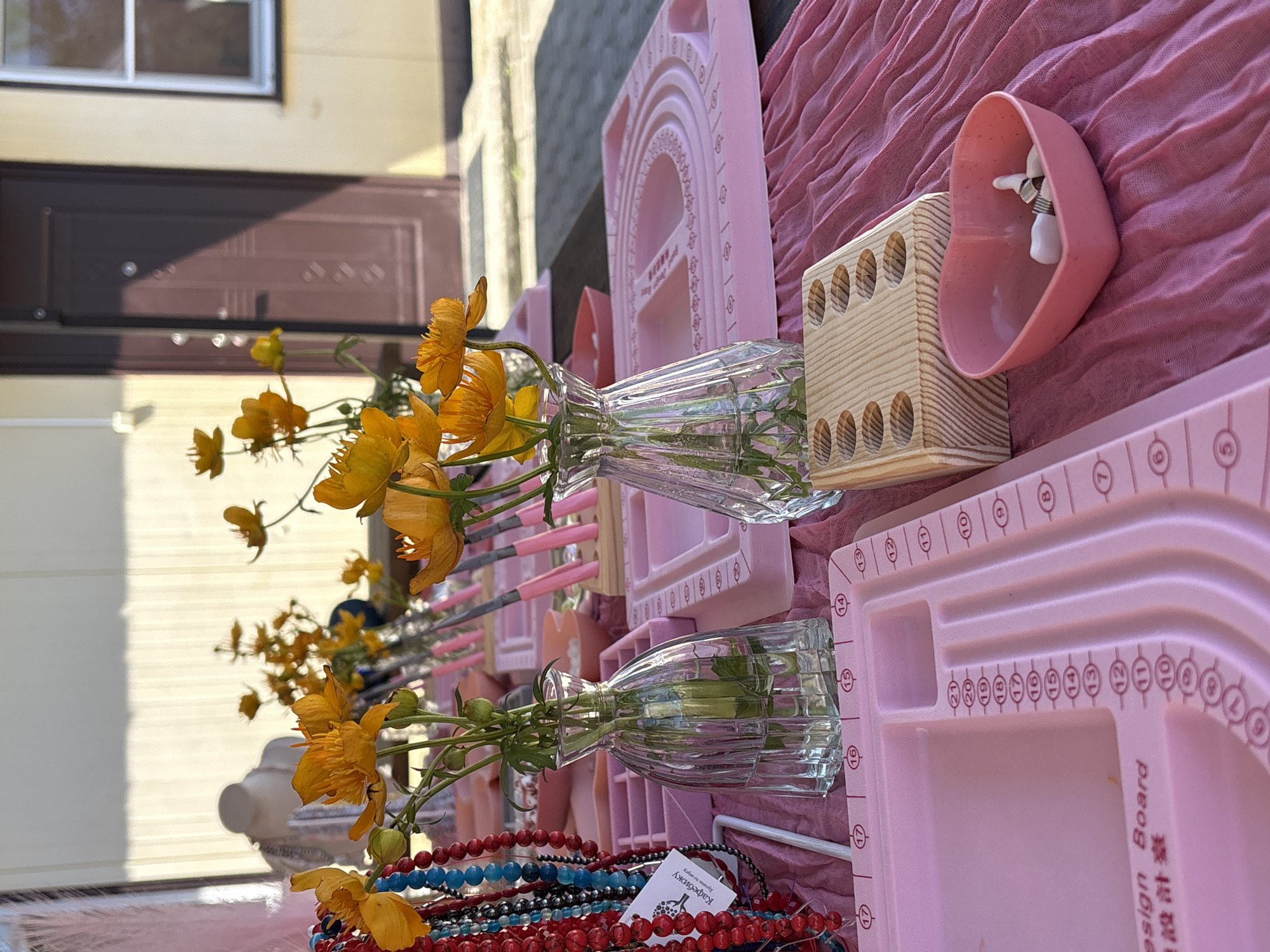 The image depicts an outdoor market stall with a focus on jewelry-making supplies. In the foreground, a pink fabric drapes across a table, showcasing various items. Two clear glass vases hold bright orange flowers, with one vase positioned next to a wooden organizer with several circular compartments. A small pink heart-shaped bowl contains what appear to be jewelry findings. To the left, a collection of colorful beaded necklaces hangs prominently, with a tag that reads "Кадребижу" in Cyrillic, along with some smaller text. Several pink jewelry design boards with numerical markings are visible, one in the lower left corner with the text "Design Board" and Chinese characters. In the background, more flowers in vases and pink jewelry design boards are arranged. A mannequin bust is partially visible on the left, adorned with jewelry. The setting appears to be in front of a building with a light-colored facade and a dark brown door with horizontal panels and a circular decorative element. A window with a white frame is also visible on the building. The lighting suggests a sunny day. While the image does not explicitly show the city of Birobidzhan, Russia, the presence of Cyrillic script on the tag suggests a Russian-speaking region. The overall scene conveys a creative and organized display for crafting and selling handmade jewelry.