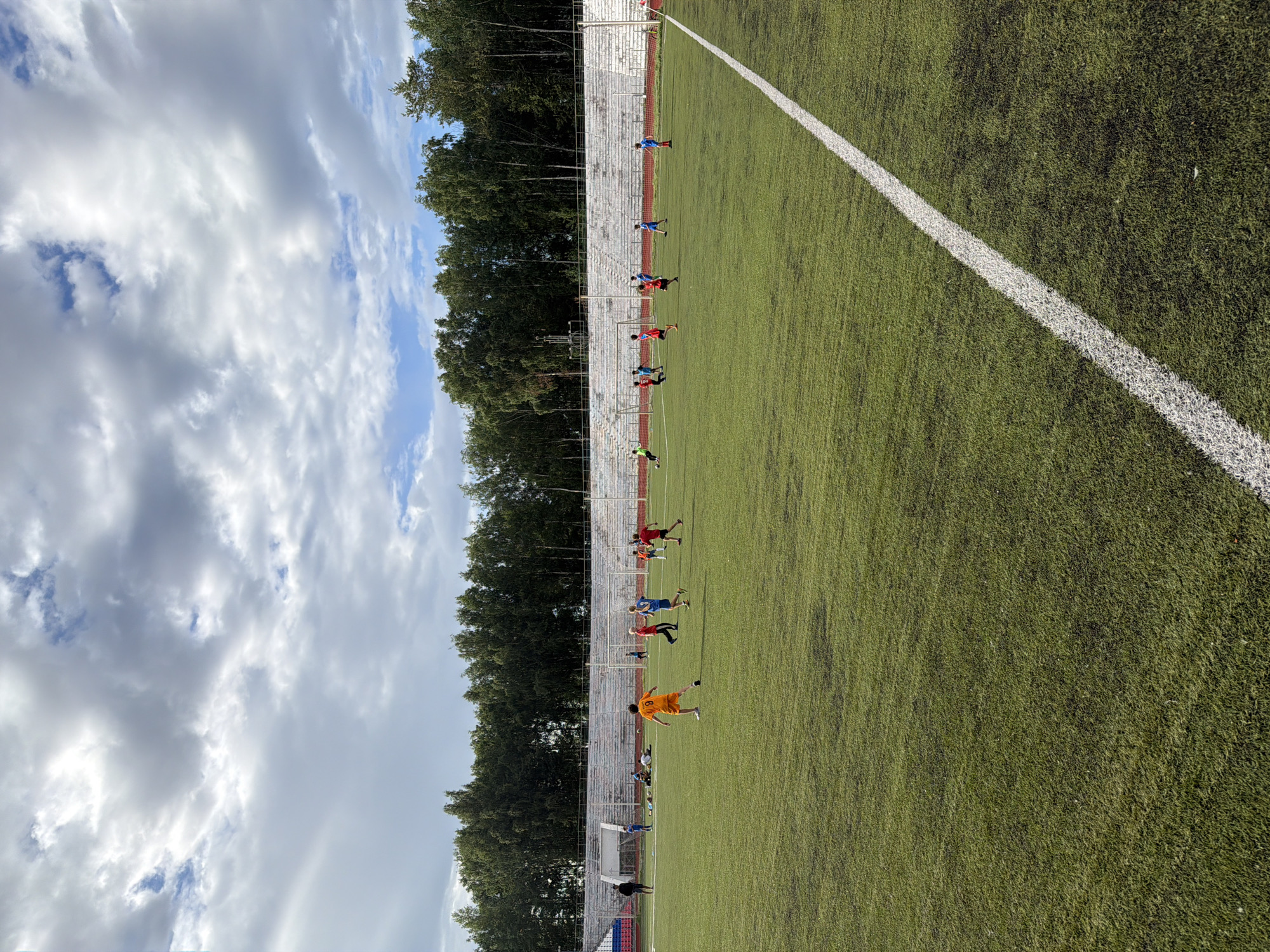 This media file captures a group of young athletes participating in a soccer practice on an outdoor artificial turf field. The scene is set in Chaikovsky, Russia, indicated by the location context. The field is marked with a white line running diagonally across the frame. Behind the field, a line of trees forms a dense green backdrop, with a grandstand of white seating visible along the left side.
The sky is a mix of blue with scattered white clouds, suggesting it is daytime, likely late morning or afternoon, with moderately bright conditions. The weather appears to be pleasant for outdoor activity.
Several individuals, presumed to be boys, are scattered across the field, engaged in various soccer-related actions. Some are in motion, running or kicking a ball, while others appear to be in a more stationary position, perhaps awaiting instruction or a pass. They are dressed in athletic wear, with some wearing brightly colored jerseys in red, blue, and orange, and one individual in an orange jersey is clearly visible in the foreground, appearing to be in mid-action. The overall atmosphere seems to be one of focused training and energetic play.
There is no visible text within the frame of the image. The composition, with the diagonal white line and the perspective from the field, draws attention to the activity of the soccer players.