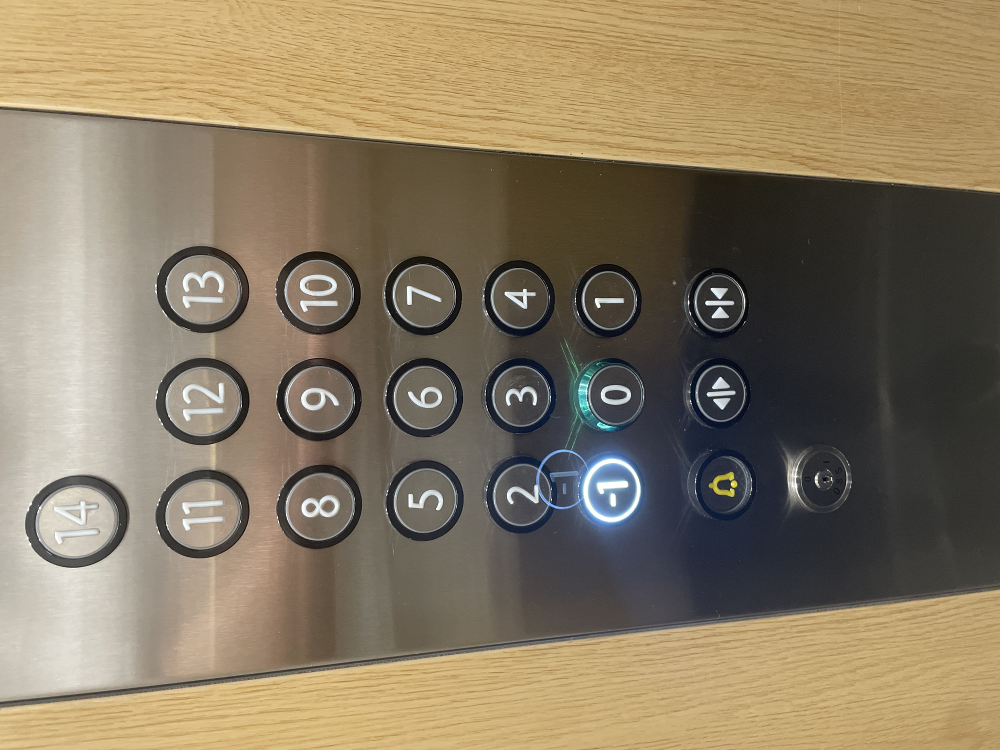 The image shows an elevator control panel with numbered buttons. The buttons are arranged in rows and columns and are made of brushed metal. The numbers range from 1 to 14, with some buttons for floors below ground level also visible, including "-1" which is illuminated, indicating it is the current floor. There are also buttons for alarm, door open, and door close. The panel is surrounded by a wooden frame. The image is taken from a slightly elevated angle, looking down at the panel. The lighting suggests an indoor environment. There are no people visible in the image. The overall scene is a close-up of an elevator control panel. There is no information in the image that would allow for the determination of its location in the city of Yunus Emre, Turkey.