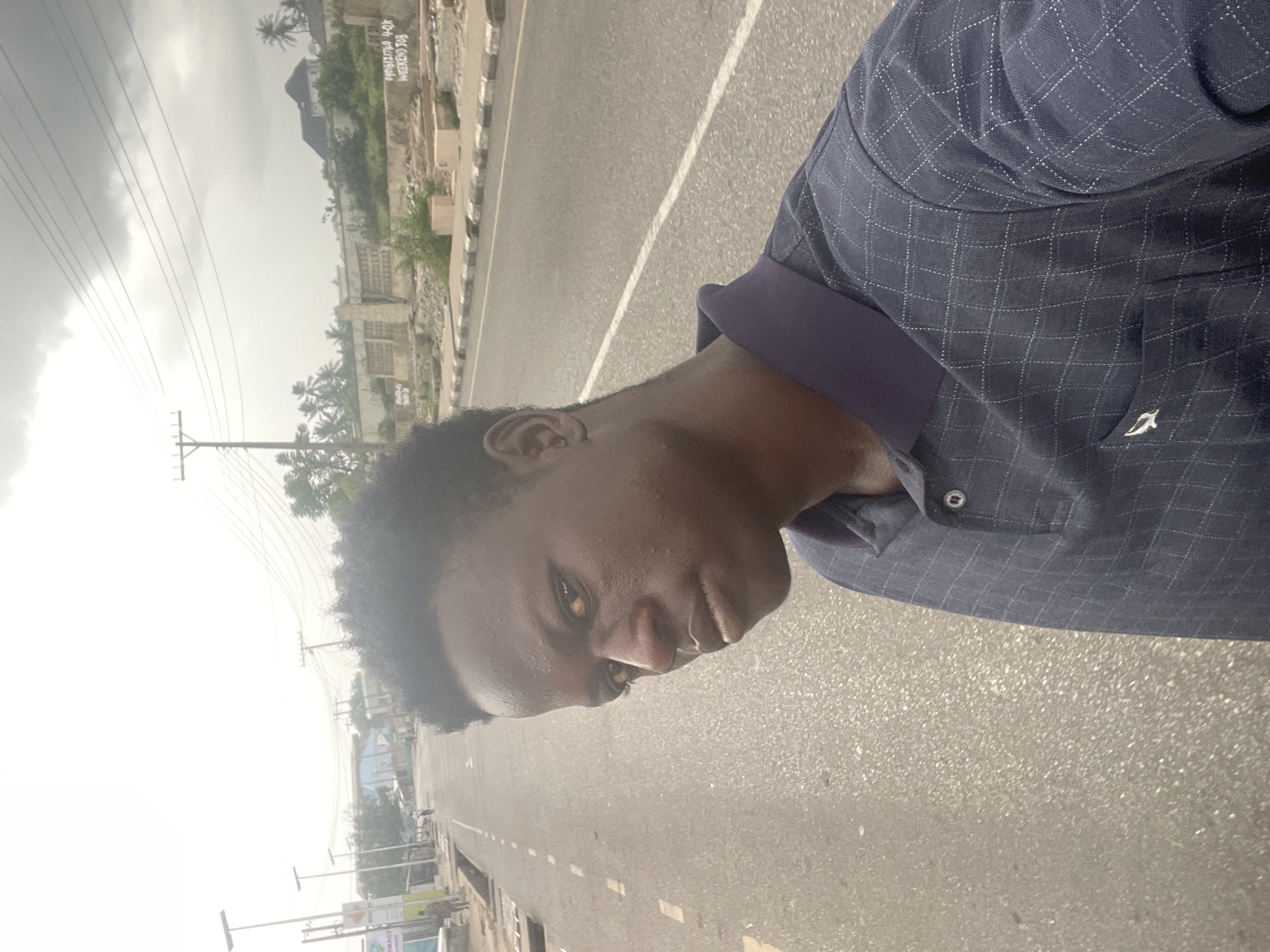 A vertical selfie features a young Black man outdoors in Badagry, Nigeria. He is positioned in the foreground, looking directly at the camera with a neutral expression. He wears a dark blue, collared shirt with a subtle light-colored checkered pattern and a small white, bird-like emblem embroidered on the left side of the chest. His skin appears slightly shiny, possibly from warmth or humidity.
The background shows a paved road with white dashed lines stretching into the distance. Utility poles and power lines are visible overhead on the left side of the frame. Along the road, there are various buildings, some appearing residential or commercial, interspersed with palm trees and other green vegetation. The sky is overcast or hazy, suggesting it is daytime.
In the upper-left background, on a distant wall or structure, partially legible text includes "BOL (WEEKEND) JOB". Further down on the left, a sign with blurry text appears to say "PHARMACY" and "ASSOCIATES". The overall scene depicts a common street environment in an urban or semi-urban area.