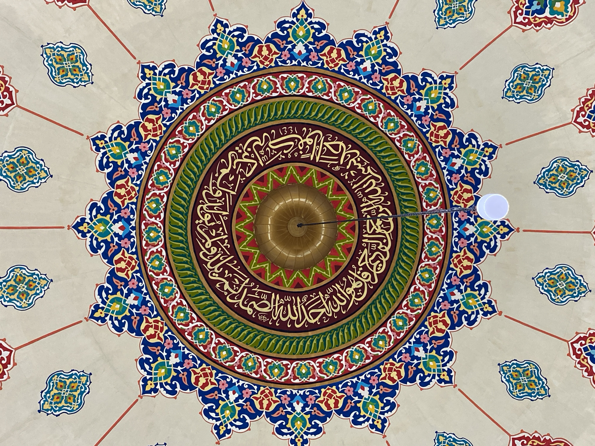 An interior, overhead shot shows the elaborate geometric and calligraphic ceiling of the Islamic Center of Lhokseumawe, Indonesia. The circular design is rich with vibrant colors, including blues, reds, yellows, greens, and gold. Concentric rings of intricate patterns, floral motifs, and Arabic calligraphy adorn the ceiling. At the very center, a brass-colored fixture is suspended, with a white light bulb hanging below it. The overall impression is one of grandeur and religious artistry.