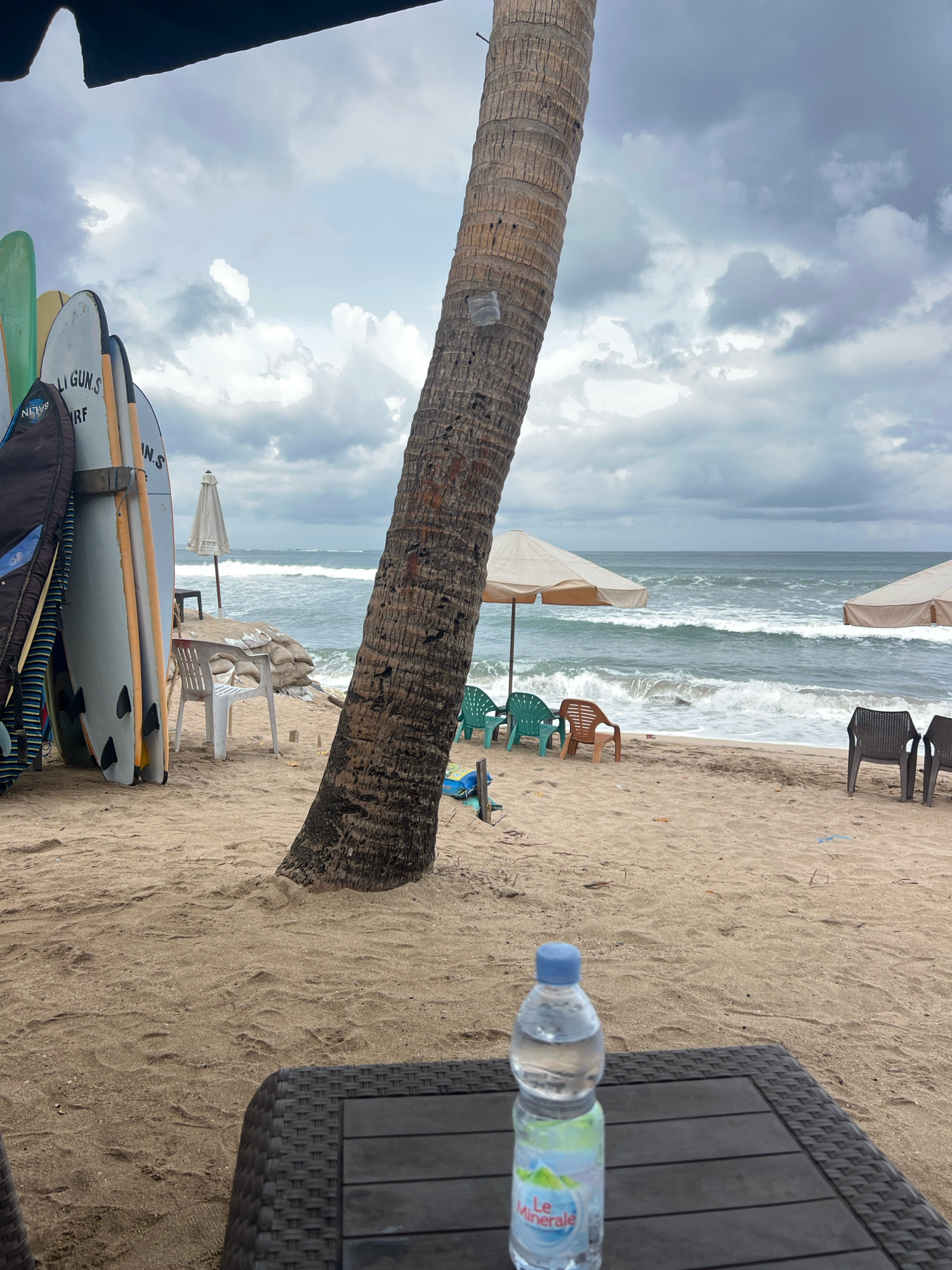 This is a medium shot of a cloudy day at a beach in Kuta, Indonesia, likely facing the ocean. In the foreground, a dark rattan coffee table holds a clear plastic bottle of "Le Minerale" water. Behind the table, a large palm tree trunk dominates the left side of the frame, with its rough bark texture visible. To the left of the palm tree, a row of surfboards are leaning against each other, some with colorful designs and brand names like "BALIN SURF" and "GUN.S".
The middle ground shows a sandy beach with several plastic chairs scattered around. Two large beach umbrellas, one beige and one white, provide shade. The ocean is visible beyond the beach, with moderate waves crashing onto the shore under an overcast sky filled with grey clouds. In the distance, a faint horizon line separates the sea from the sky. Sandbags are piled near the water's edge, and some debris is visible on the sand. There are no people or animals in the visible scene.