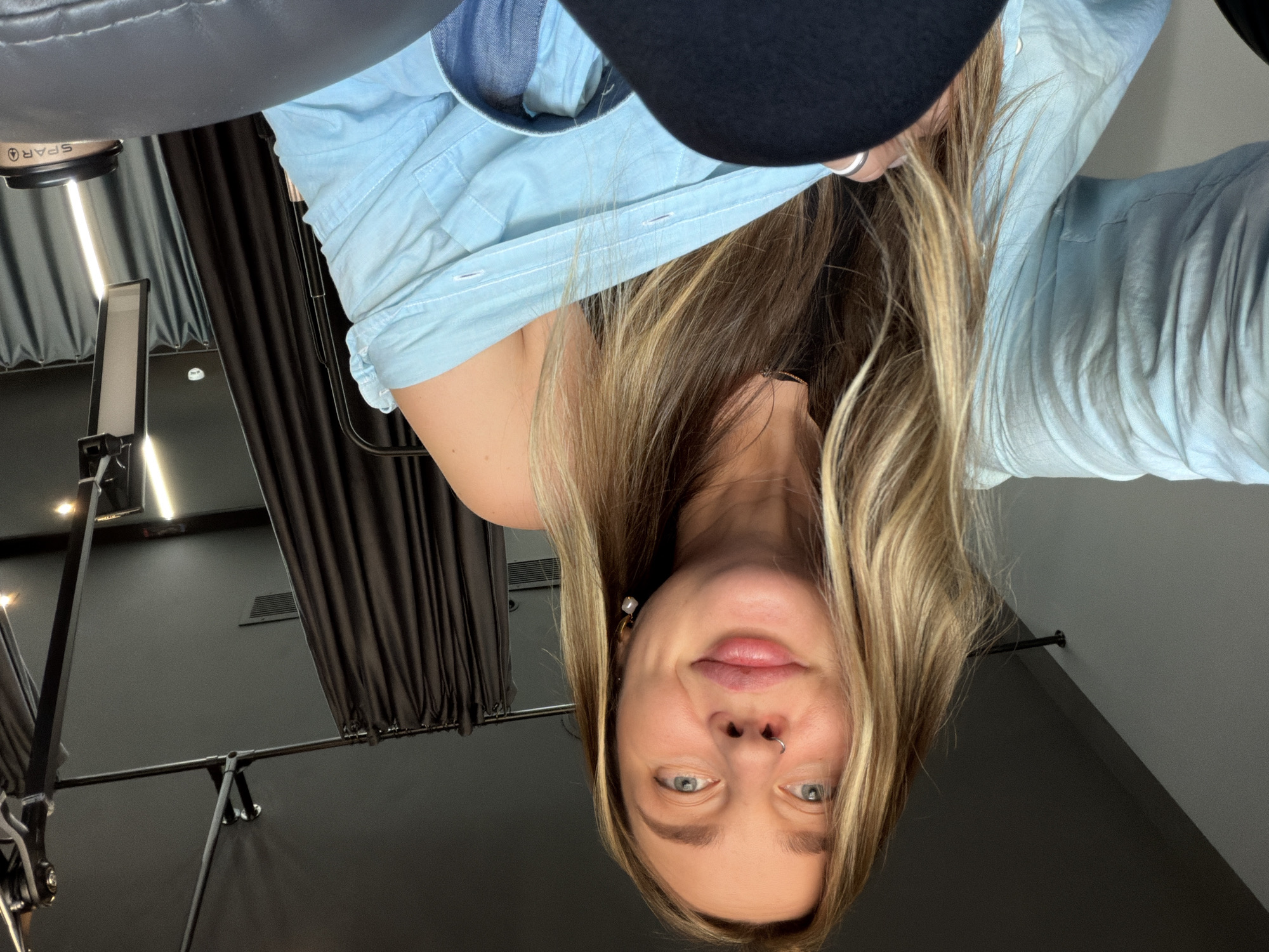 The image is a selfie taken from a high angle, featuring a woman with blonde hair wearing a light blue shirt and a black garment visible at the top. She has light eyes and a nose ring. The background includes dark drapes, a lighting setup, and a black wall. The image appears to be taken indoors, possibly in a studio setting. There is no information in the image to suggest it was taken in Moscow, Russia.