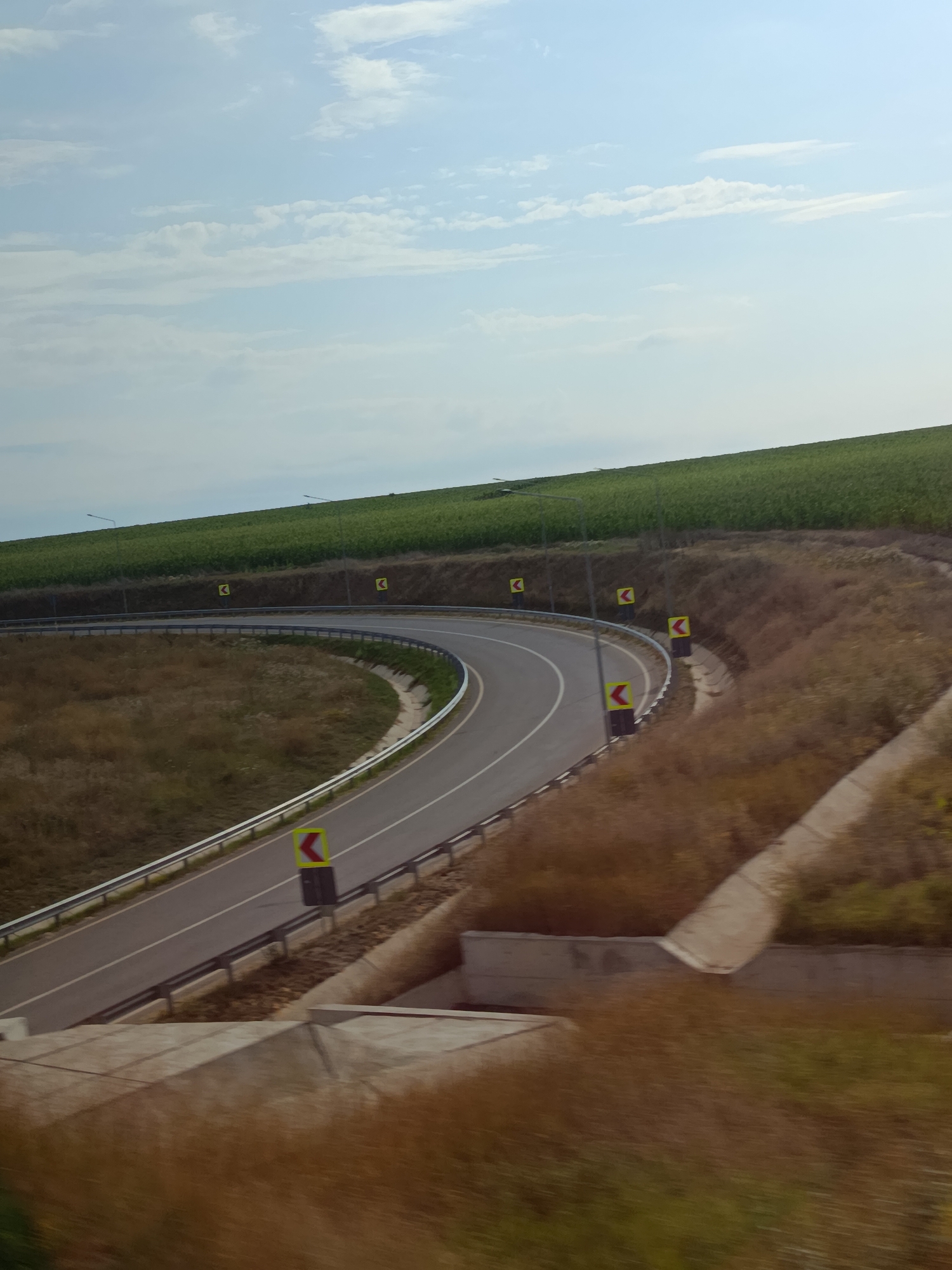 The image captures a curved asphalt road with white lane markings, bordered by a metal guardrail. Several yellow diamond-shaped warning signs with black arrows indicating curves are placed along the roadside. The road is surrounded by dry, brown vegetation and a concrete drainage channel. In the background, a steep green field, possibly a cultivated crop, rises towards a pale blue sky with scattered white clouds. A few streetlights are visible on poles atop the green field. The scene appears to be on the outskirts of Comrat, Moldova, on a clear day.