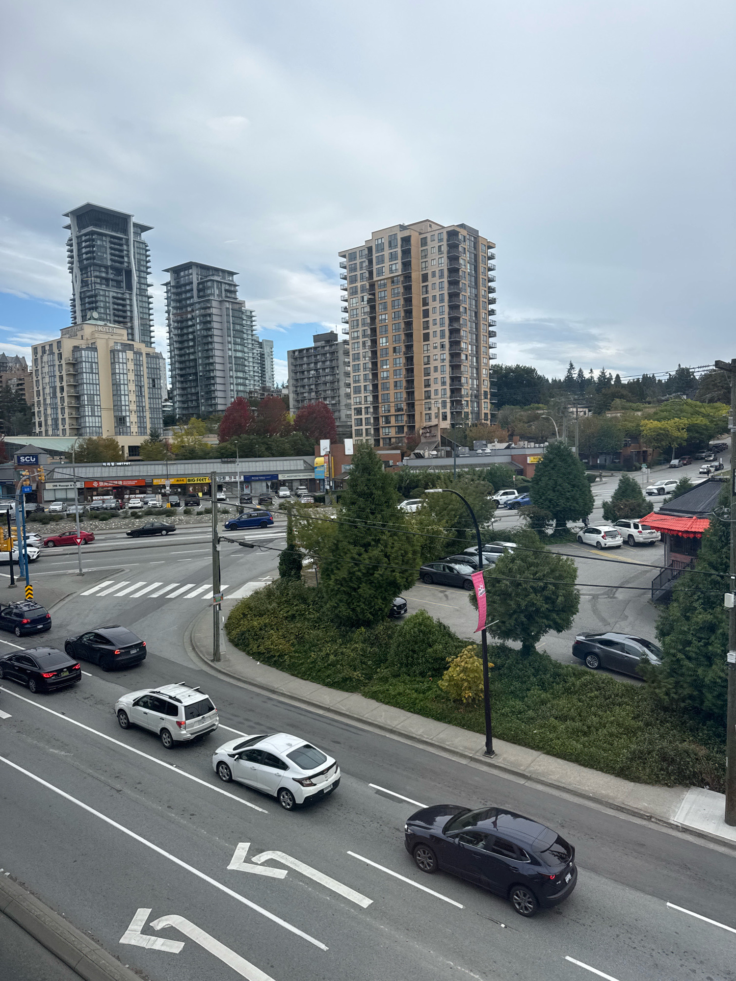 The image shows a street scene in Burnaby, Canada, likely near the intersection of Kingsway and Edmonds Street, a busy commercial and residential area. Several multi-story buildings, including apartment complexes and commercial establishments, are visible in the background, with the distinctive towers of "The Sovereign" residential complex prominent to the left. The foreground displays a multi-lane road with traffic. Cars are in motion and parked. One white SUV is parked to the right of the road, next to a building with a red roof. A dark-colored sedan is driving in the foreground on the right lane. Several other cars are on the road, including a white sedan in the middle lane and a black sedan on the far left. The sky is overcast, suggesting a cloudy day. Greenery, including trees and bushes, lines the sides of the road and parking areas. A pedestrian crossing is visible on the road. Buildings in the middle ground appear to house various businesses, including restaurants like "BIG FEET" and a "SCU" (likely Simon Fraser University Credit Union). The overall scene captures a typical urban environment in Burnaby, characterized by a mix of residential and commercial development.