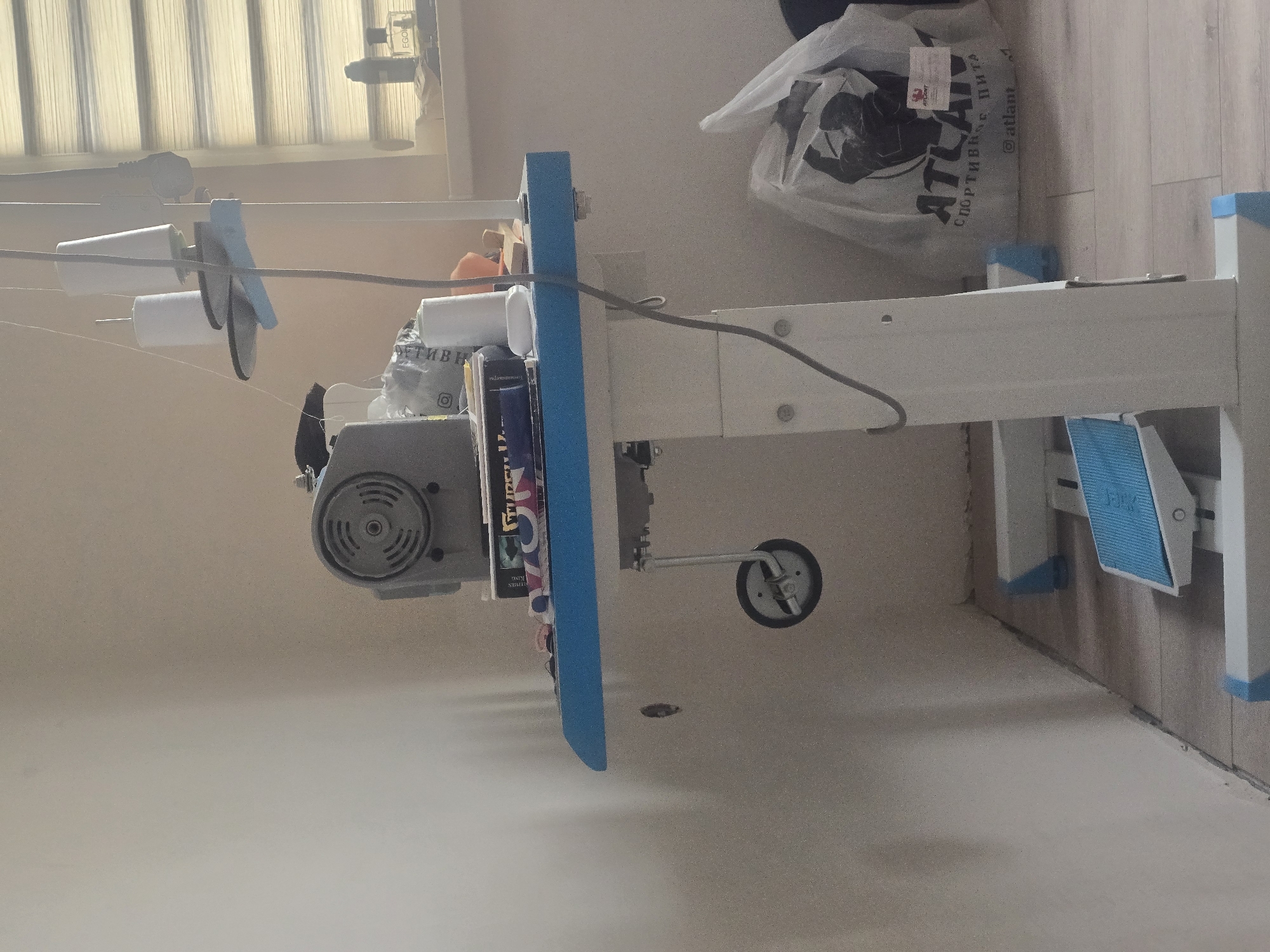 The image depicts an industrial sewing machine with a white metal stand, situated in a room. The sewing machine unit itself is gray and mounted on a blue-edged table. Several spools of thread are visible, along with various items on the table, including what appear to be books or binders. A foot pedal for the sewing machine is visible on the floor. To the right of the sewing machine, a large white plastic bag with a graphic design and Russian text is present. In the background, a window with horizontal blinds is visible. The walls are painted a light color, and the floor appears to be wood or laminate. The lighting suggests it is daytime, with light coming from the window. There are no people visible in the image. The scene appears to be an indoor workspace. The location is not specified within the image.