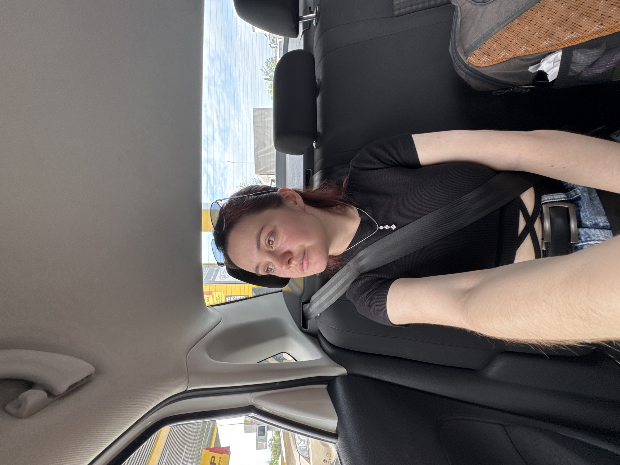 A woman with auburn hair and sunglasses is seated in the back of a car, wearing a black top and a seatbelt. A black backpack is on the seat next to her. The car is passing a commercial area with buildings and parking lots, one building having yellow signage with Cyrillic letters possibly indicating the location is in Sochi, Russia. The sky is partly cloudy.