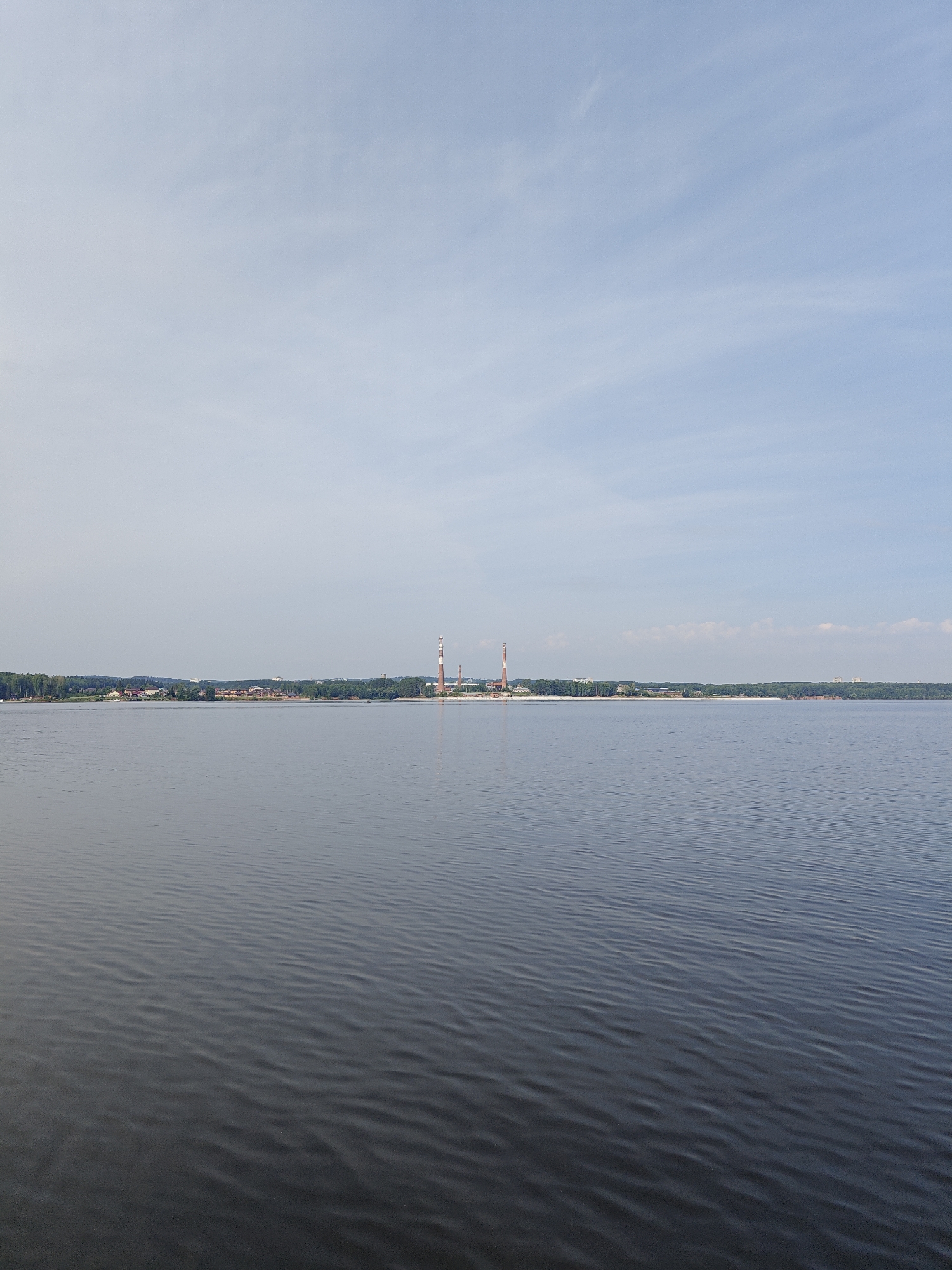 The image shows a wide, calm body of water, likely a river or reservoir, in Chaykovsky, Russia. The water occupies the majority of the lower half of the frame, with gentle ripples visible. Across the water, a distant shoreline is lined with trees and buildings, suggesting a populated area. Prominently on the right side of the distant shore, there are two tall, industrial smokestacks, characteristic of a power plant or factory. The sky above is a pale blue with scattered, thin clouds, indicating fair weather. The scene is serene and expansive, with no people or boats immediately visible on the water.