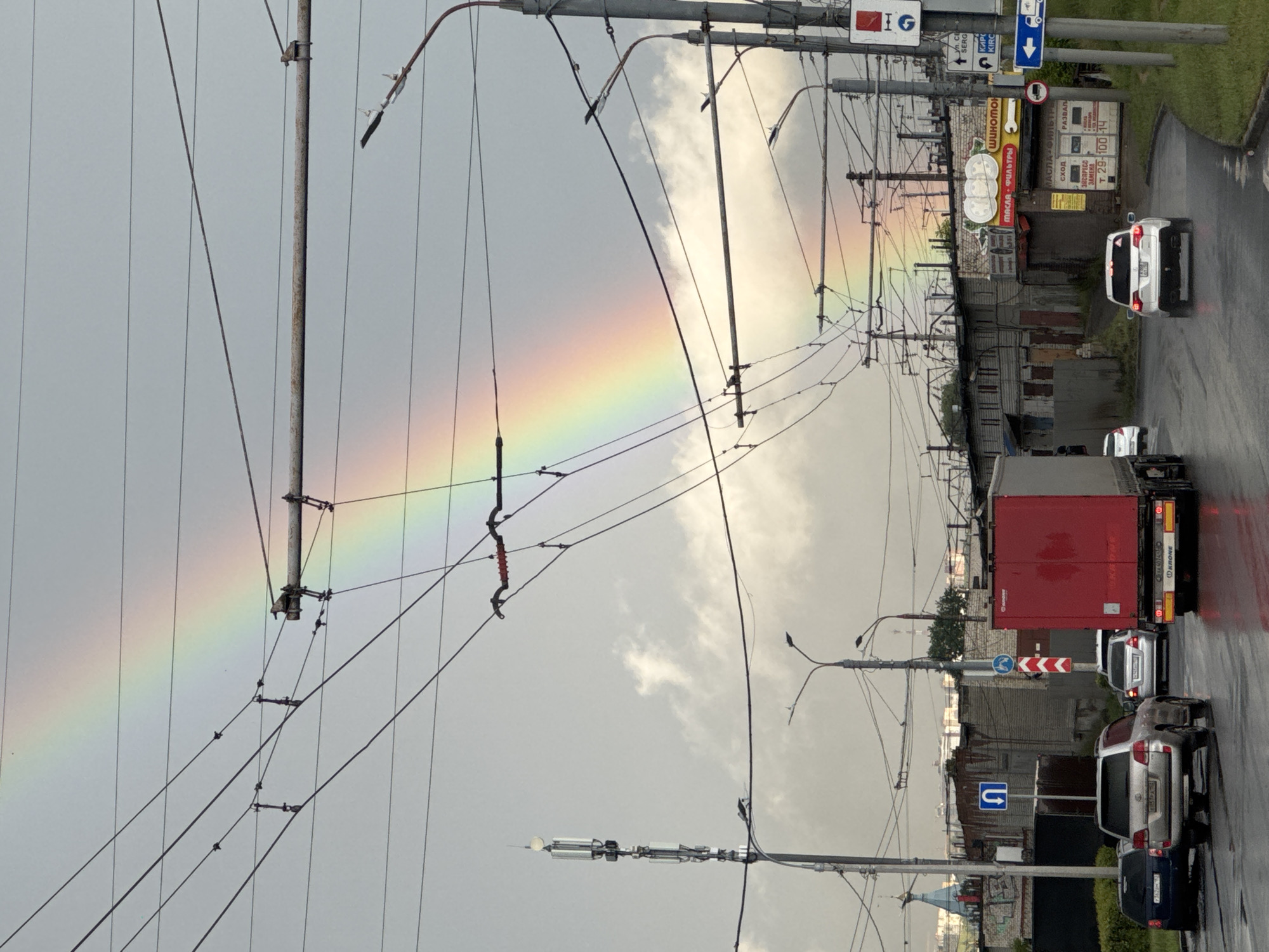 The image shows a street scene in Nizhny Novgorod, Russia, with a prominent rainbow arcing across a cloudy sky. Power lines and street lights are visible, indicating an urban environment. On the street, several vehicles are present, including cars and a red truck. Buildings line the street, some appearing to be industrial or commercial, with signs visible in Russian. The ground is wet, suggesting recent rain. A cell tower is also visible on the left side of the image.