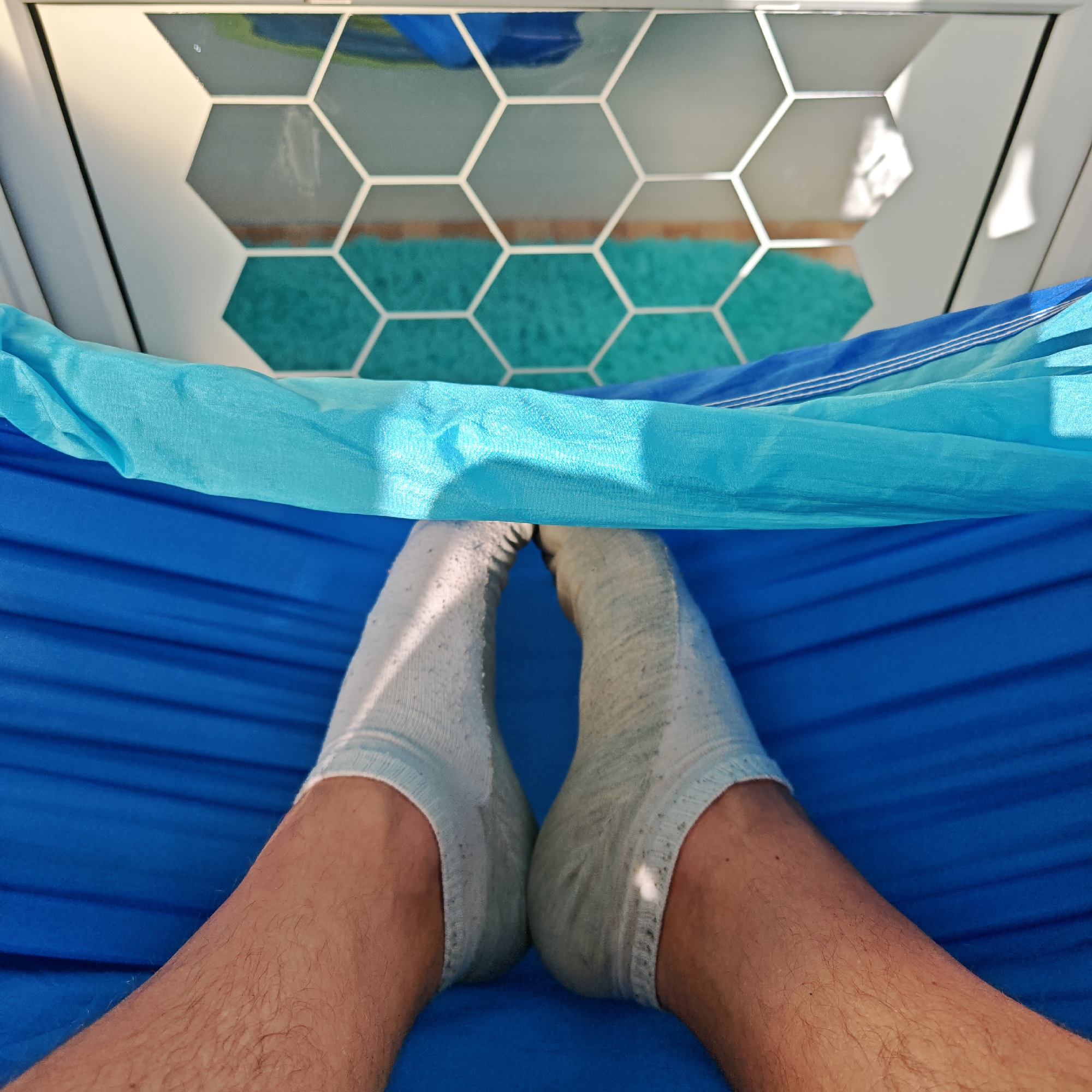 A first-person perspective shot features a person reclining in a blue hammock, their feet clad in light-colored socks, resting within the hammock's confines. A section of light blue fabric is draped overhead. Above the hammock, a window with a white frame and a honeycomb pattern is visible, reflecting a turquoise rug and blue objects. The specific location within Rostov, Russia, is not discernible from the image.