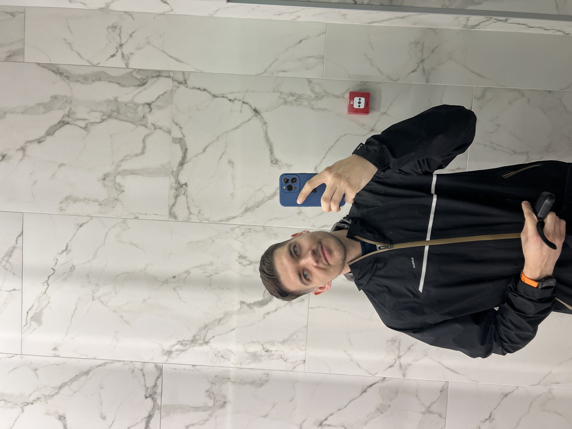 A male individual is captured in a mirror selfie indoors within Kazan, Russia. He has short brown hair, blue eyes, and light stubble, looking upwards and to his right towards the camera. He wears a black hooded jacket with a brown central zipper and a prominent white horizontal stripe across the upper chest. The mirrored text "KIPRUN" is visible above this stripe. An orange-strapped device, likely a smartwatch or fitness tracker, is on his left wrist. He holds a blue-cased smartphone (with a visible triple-lens camera array) in his right hand, pointed towards himself, and a small, black, cylindrical object in his left hand. The background wall is covered in large white tiles featuring grey marble-like patterns. A red fire alarm call point, marked with a white symbol of four circles surrounding a central dot, is mounted on the wall to the right of the subject.