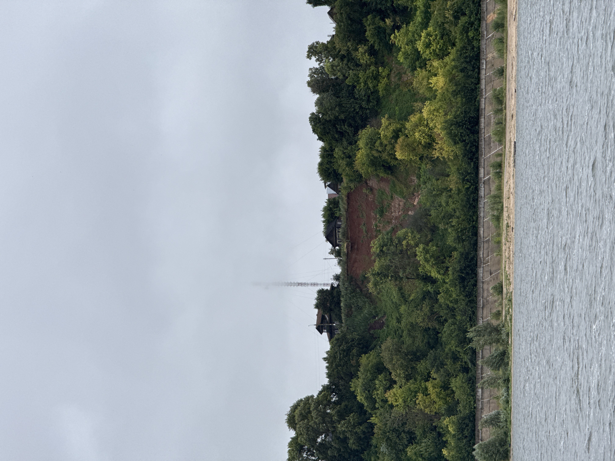 A wide shot captures a grey, overcast sky above a treed embankment and a body of water. The embankment, sloping upwards from the water, is covered in lush green trees. A red-brown scar indicates a landslide on the embankment. Several houses and a tall, lattice telecommunications tower are visible on top of the embankment. A concrete wall with diagonal supports runs along the base of the embankment, separating it from a sandy shore and the water. The water in the foreground is choppy, with visible ripples and waves. The scene is likely a view of the Volga River in Kozmodemyansk, Russia.