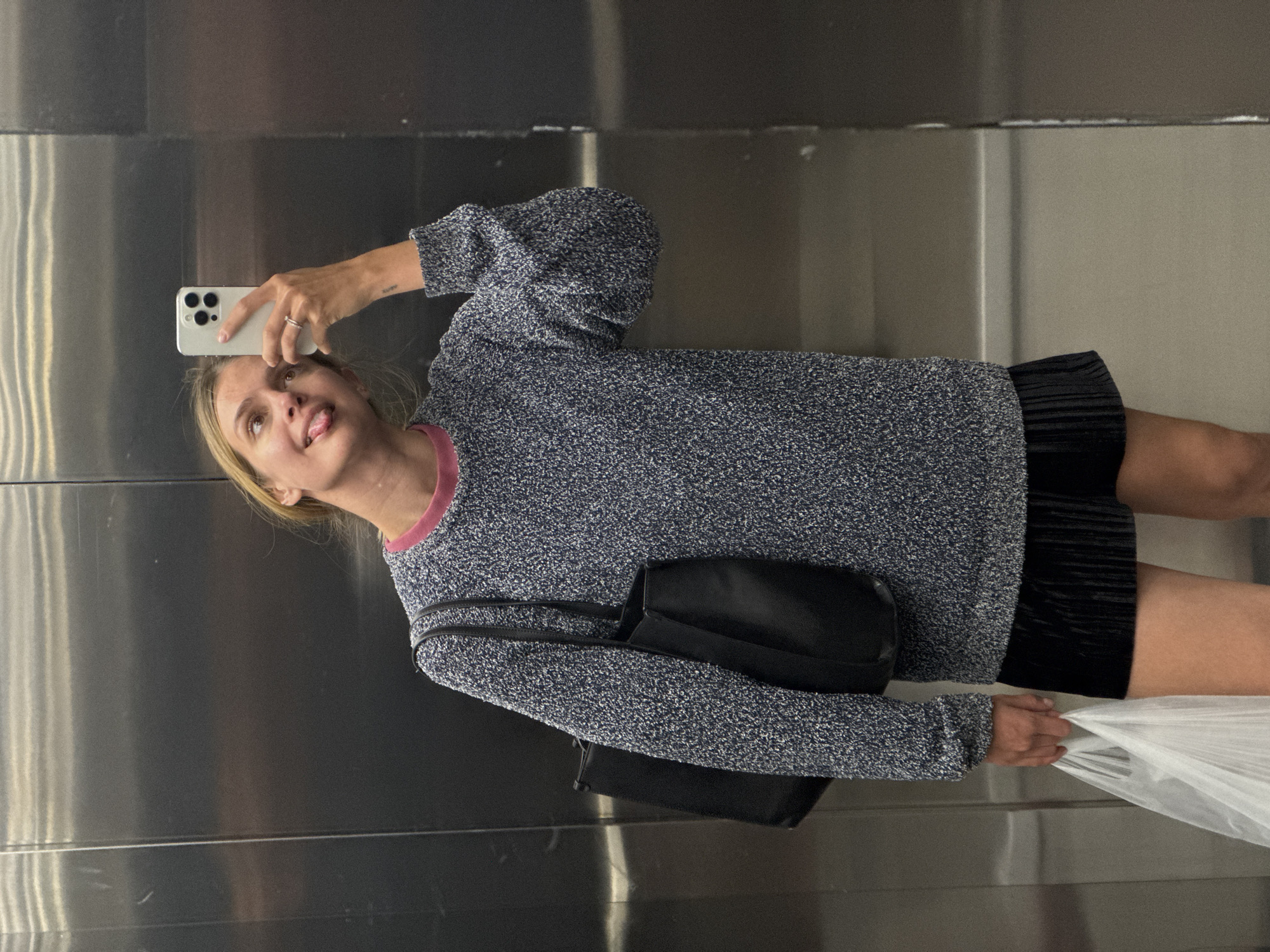 A fair-skinned woman is taking a selfie with a white smartphone while standing in an elevator. She is wearing a speckled blue and white sweater with a pink collar, a black skirt, and a black over-the-shoulder bag. She is holding a white plastic bag in her right hand, which is on the right side of the frame. The woman's tongue is sticking out as she looks up at the phone. The elevator is made of stainless steel with horizontal seams. The location is in the city of Unknown, Unknown.
