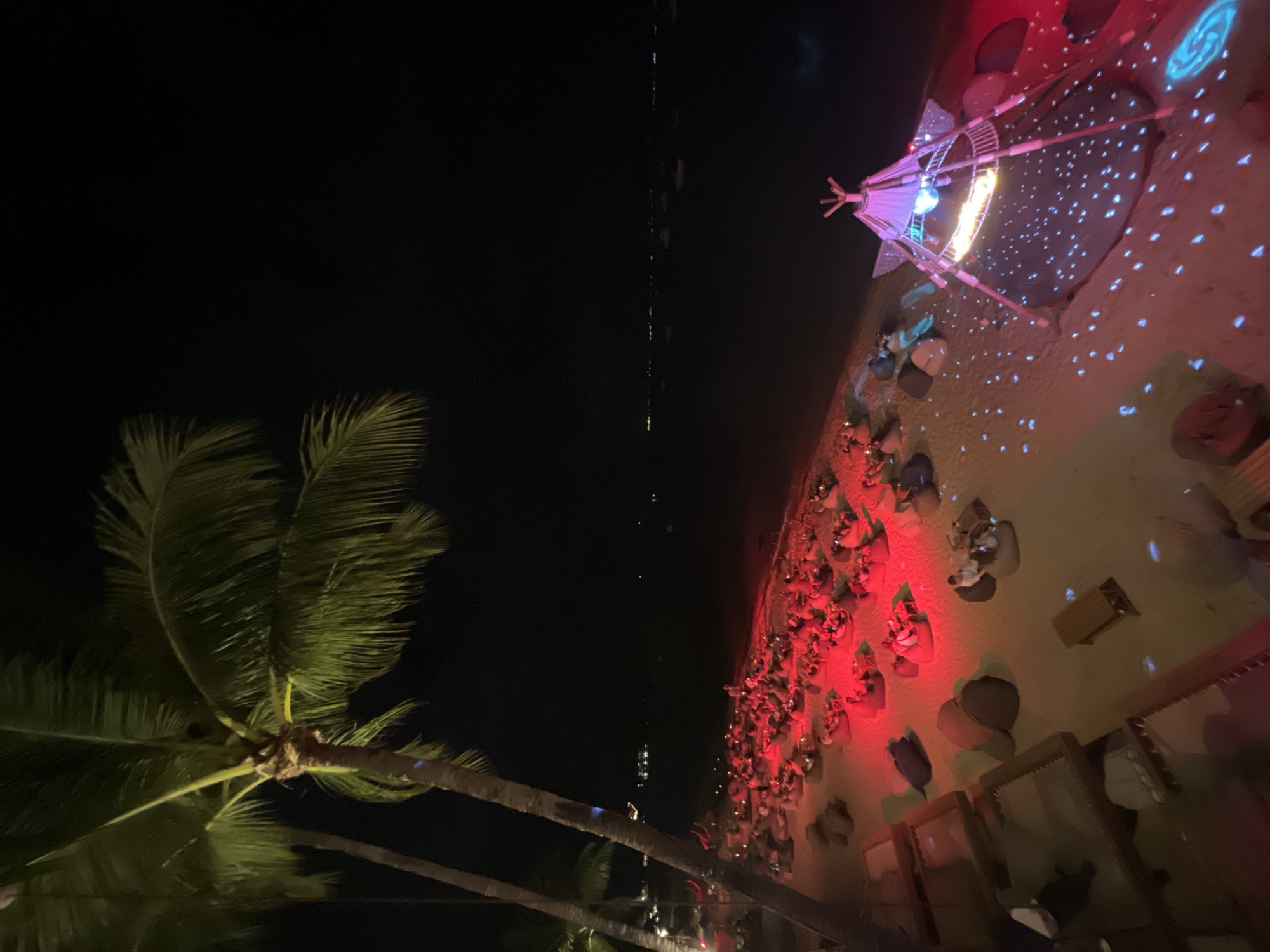 The image depicts a nighttime scene on a sandy beach in Baan Bo Phut, Thailand. In the foreground, several palm trees with green fronds stand tall against a dark sky. A beach area is set up with numerous red-lit seating areas, each featuring small tables and beanbag chairs. Several people are visible, seated and interacting at these tables. To the right, a decorative tent structure with colorful lighting and a disco ball inside is set up on the sand, with scattered blue lights projected onto the ground around it. The sea stretches out in the background, dotted with faint lights from distant boats or structures. The overall atmosphere suggests an outdoor dining or entertainment venue.