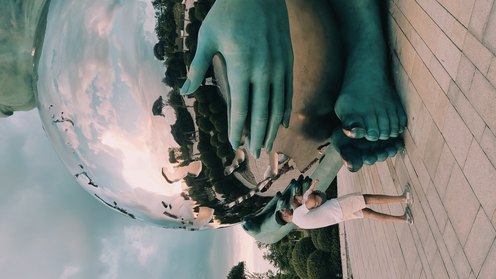 A man holds a baby while standing on a paved area in front of a large, metallic, spherical sculpture with reflective surfaces, which is supported by colossal teal-colored hands and feet. The sculpture is situated outdoors, with manicured hedges and trees visible in the background, along with scattered clouds in the sky. The scene appears to be in a public park or sculpture garden. The man is wearing a white t-shirt and light-colored shorts, and the baby is held in his arms. The sculpture's reflective surface mirrors the sky and the surrounding landscape. This sculpture is located in Krasnodar, Russia, specifically within the "Solnechny Ostrov" (Sunny Island) park.