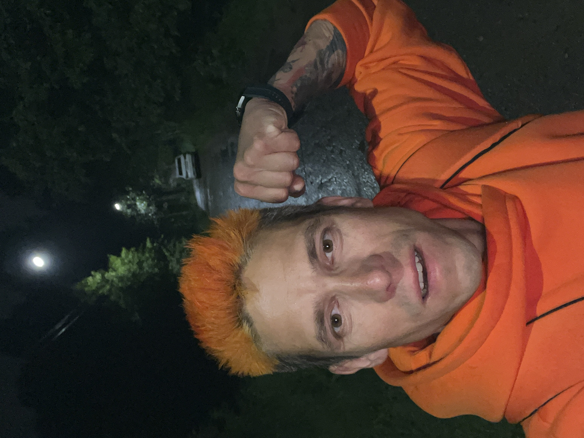 This image, captured in Chaikovsky, Russia, presents an adult male posing outdoors at night. The man, with bright orange, spiky hair and brown eyes, faces the camera with an intense, determined expression, his mouth slightly agape revealing some lower teeth, and his nose appearing slightly flushed. He wears an orange hooded sweatshirt featuring dark trim lines. His left arm is flexed, with his hand clenched into a fist, and a dark smartwatch is visible on his wrist. Intricate tattoos are discernible on his left forearm. The background shows a dark, outdoor scene with a wet, paved path or road receding into the distance, bordered by dense, dark green trees. A bright street light illuminates a section of the path and trees on the left. Further down the path, a white SUV or car is visible. Against the dark night sky in the upper left, a prominent, luminous crescent moon is visible, along with faint parallel lines suggestive of overhead power cables.