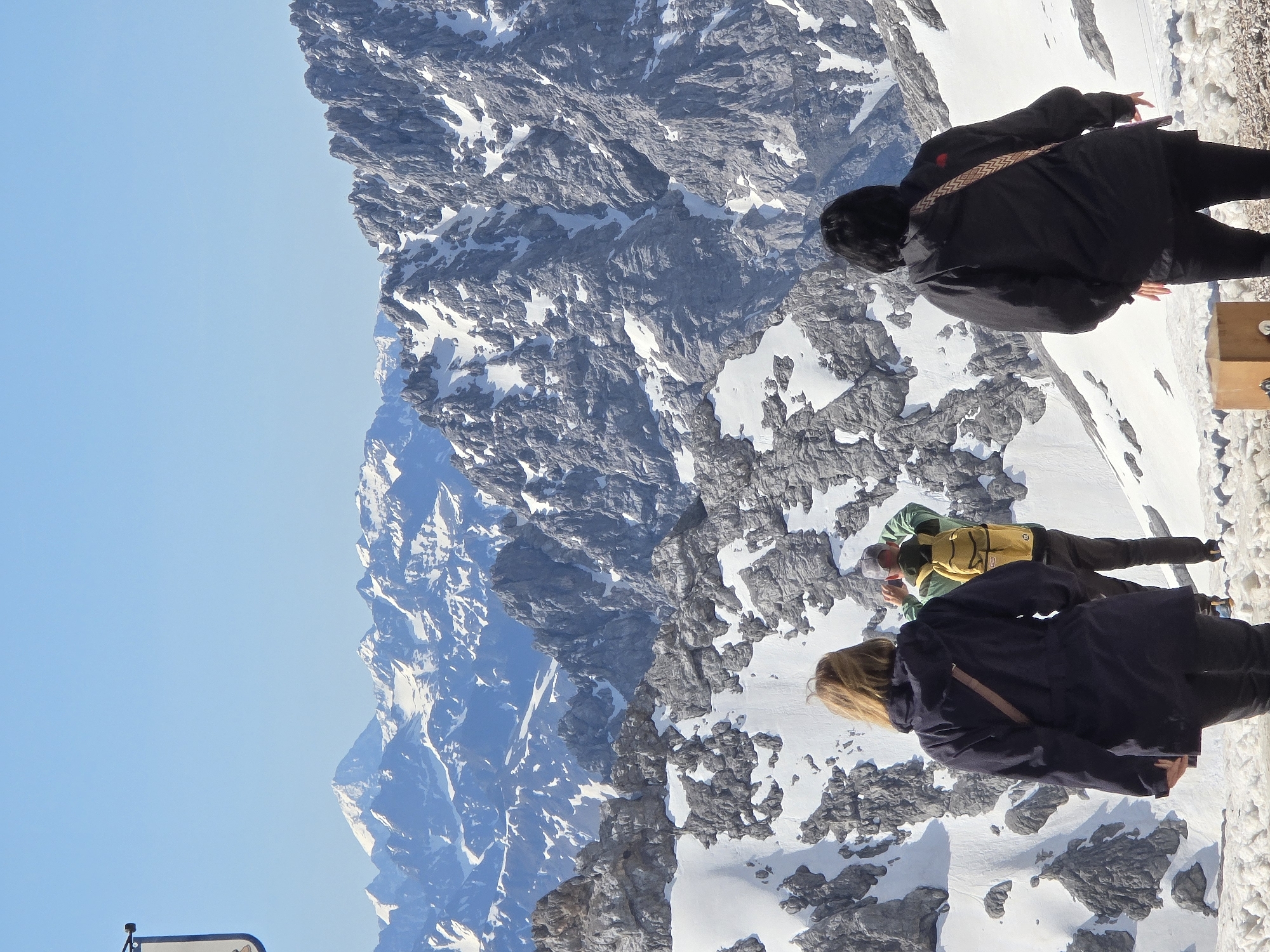The image depicts a scene in the Bavarian Alps, likely near the summit of the Zugspitze, which is accessible from Garmisch-Partenkirchen, Germany. In the foreground, three individuals are observed from behind, facing a vast, snow-covered mountain range under a clear blue sky. The closest person on the left is a woman with blonde hair, wearing a dark navy jacket and black pants. She is holding a brown strap across her shoulder. To her right, a man in a green jacket and dark pants is taking a photo with his phone, wearing a yellow backpack. Further to the right, another woman with black hair is wearing a black jacket and black pants, with a patterned strap across her shoulder. In the lower right corner, a small wooden box or pedestal is visible. The background is dominated by jagged, snow-dusted mountain peaks and slopes, indicating a high-altitude alpine environment.
