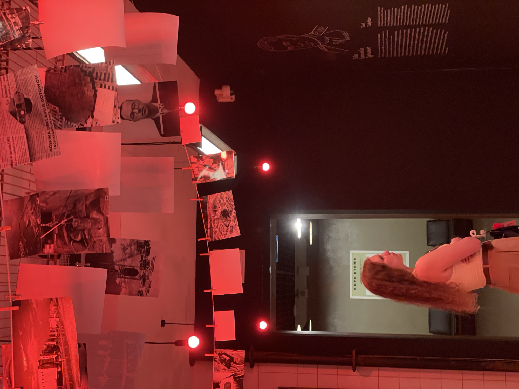 The image depicts an interior space illuminated by a reddish light. There is a display of photographs and documents attached to a metal frame, some hanging from clothespins, possibly an art installation. On the ceiling, there are drawings or illustrations. Through a window or doorway, a woman is visible, standing in another room. The image is taken in what appears to be a public space, possibly a restaurant, bar, or gallery. No specific evidence definitively places the location in Moscow, Russia.