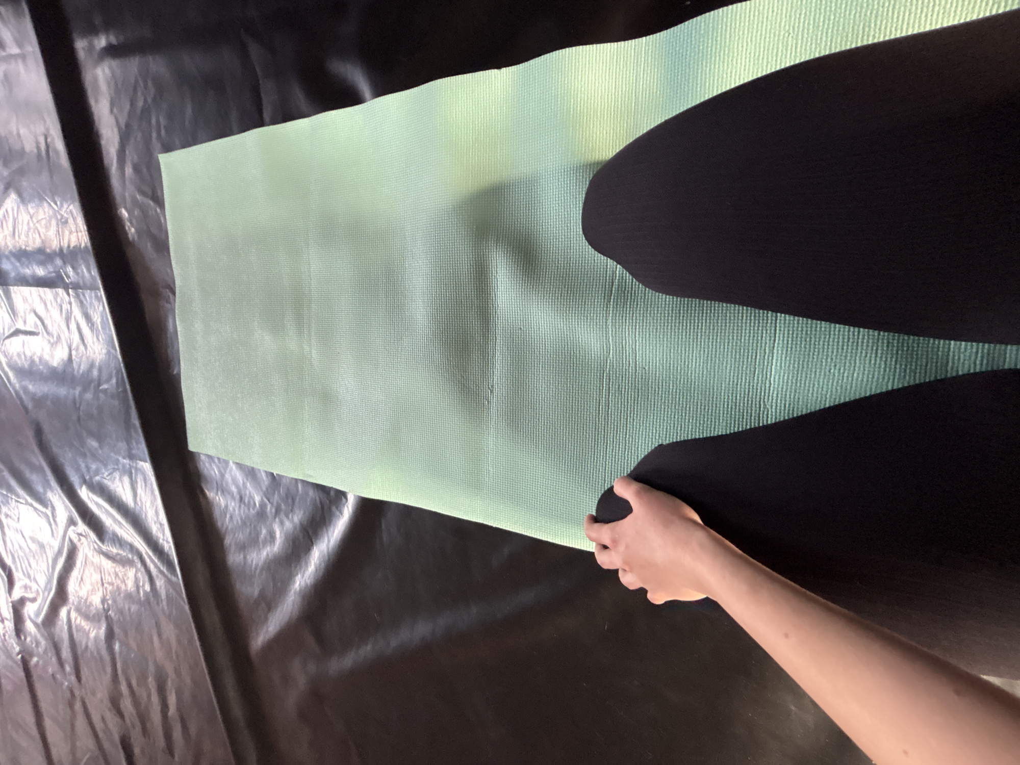 A high-angle shot reveals a light green yoga mat laid out on a black plastic sheet. A person wearing black leggings is kneeling on the mat, their left hand grasping the edge of the mat near the lower right corner of the image. The background is indistinct, but the setting appears to be indoors in Ферма, Russia.