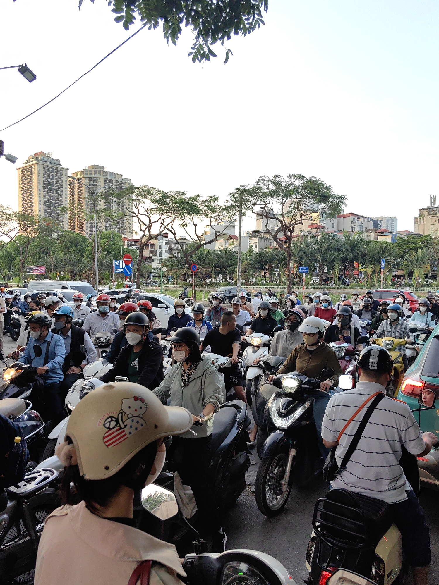 The image depicts a dense traffic scene in Hà Nội, Vietnam. A large group of people on motorbikes are densely packed together on a street. Most of the motorbike riders are wearing face masks, and some are wearing helmets. The background features buildings, trees, and traffic signs. In the foreground, one of the riders is wearing a helmet that features Hello Kitty designs. A mixture of modern and older buildings are visible, indicative of an urban area. Overall, the scene represents a typical traffic situation in a Vietnamese city.