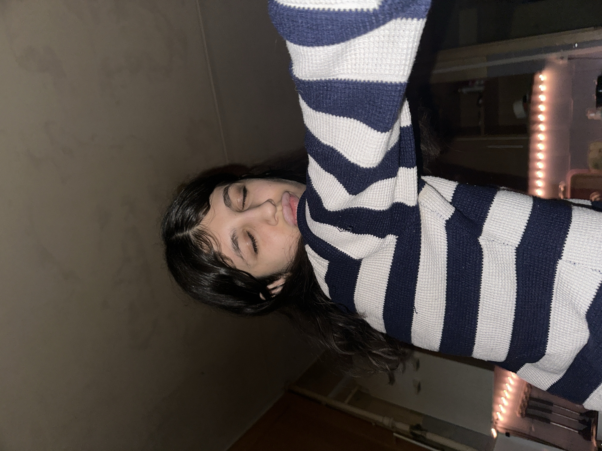 A young person is lying on their side in a room with dark walls. They are wearing a striped sweater with dark blue and white stripes. Their eyes are closed, and their lips are slightly parted. The background is dimly lit, but there appears to be a string of lights in the distance. The location is unknown, but not Moscow, Russia.