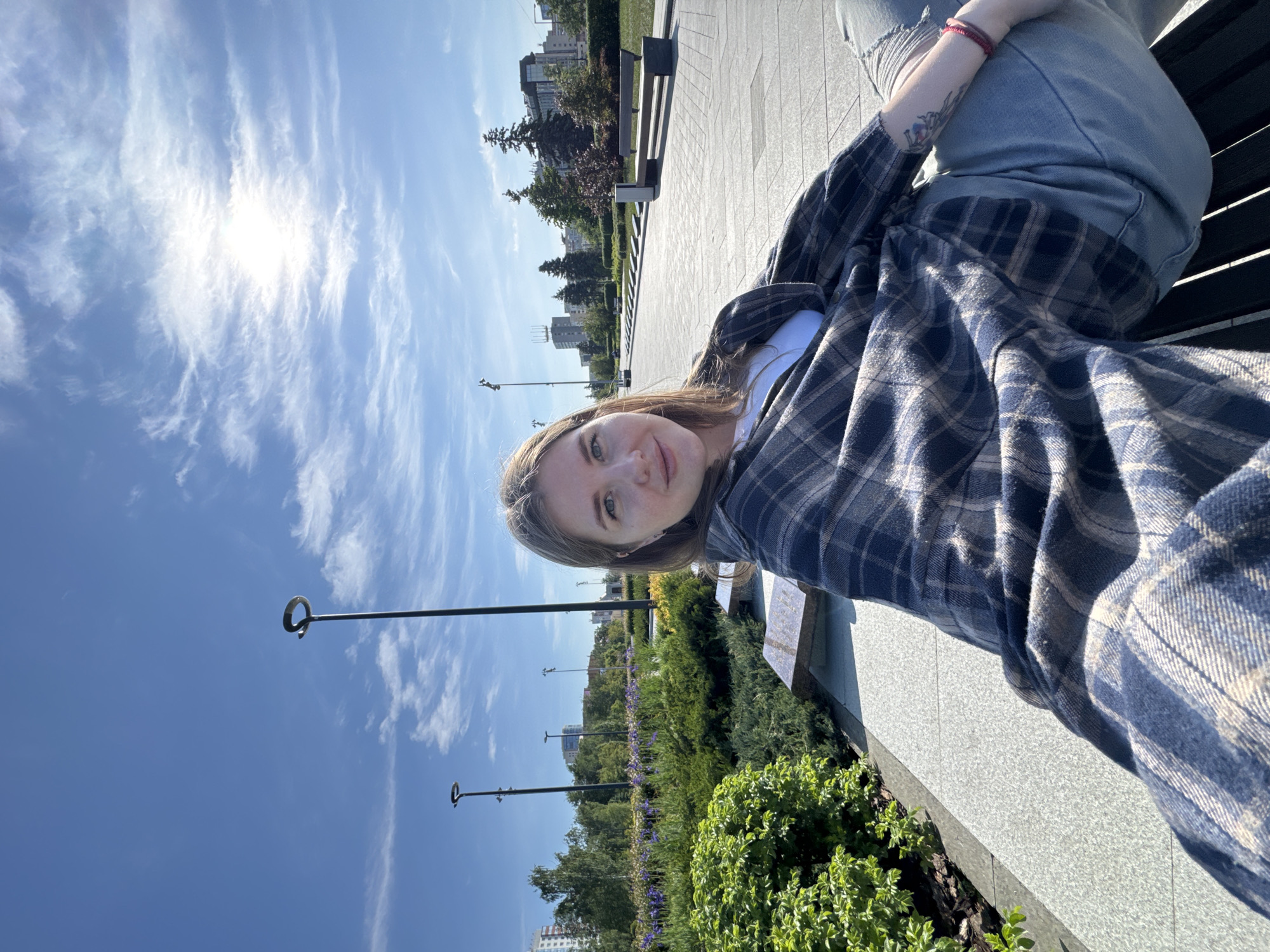A young woman is captured in a well-lit, outdoor setting, likely an urban park or promenade in Perm, Russia. The image is oriented sideways, but depicts a bright, sunny day with a clear blue sky dotted with white, wispy clouds, and the sun visible on the left side of the frame.
The woman is seated on a dark-colored bench on the right side of the image, looking directly at the camera with a slight, pleasant smile. She has light brown hair, fair skin, and green eyes. She is dressed in a blue, brown, and white plaid shirt worn over a white top, paired with light-colored jeans or trousers. A red bracelet adorns her left wrist, and a tattoo is visible on her left forearm.
To her left, a wide, paved walkway stretches into the background, flanked by manicured green bushes, trees, and some purple flowers. Several dark lampposts with circular tops line the path, receding into the distance. In the background, modern urban buildings are visible through the trees, confirming the city setting. The overall atmosphere is bright and serene, characteristic of a pleasant day out. There is no visible text in the image.