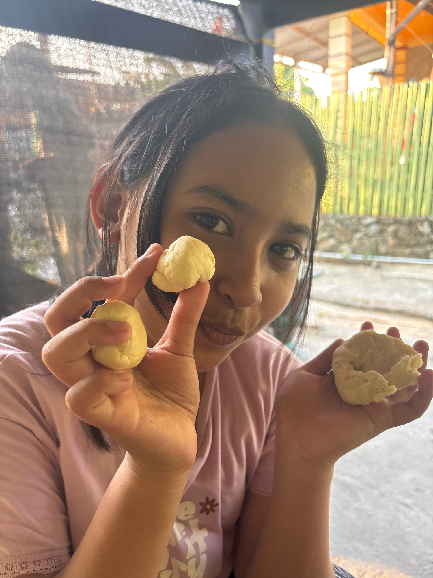 A young Indonesian female in Arateng, Indonesia, is in a close-up shot, holding several small balls of dough in her hands. She is wearing a pink t-shirt. The background shows a fence made of bamboo and some structures, suggesting an outdoor setting. The lighting indicates it is daytime, with sunlight filtering through. The image focuses on the woman's hands and face as she presents the dough.