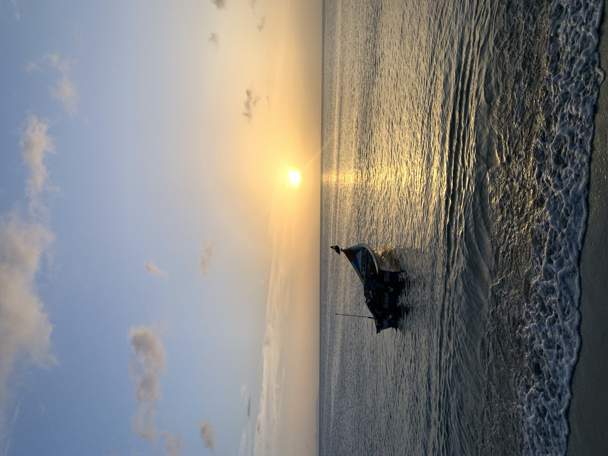 A fishing boat with an outboard motor is anchored in calm waters near the shore at sunrise. The sun, a bright yellow orb, is positioned in the upper right quadrant of the image, casting a golden reflection on the water. A bird is perched on the stern of the boat. The sky is a gradient of pale blue and yellow, with scattered clouds. The foreground shows the gentle waves of the ocean lapping against a sandy beach. The scene depicts a serene morning in Juan Griego, Venezuela.