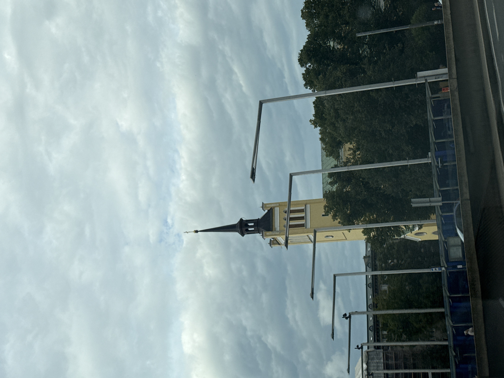 The image shows a view of Tallinn, Estonia, on a cloudy day. In the background, the spire of St. Olaf's Church (Oleviste kirik) is visible, a prominent landmark in the city. The church is a tall, slender structure with a yellow facade and a dark, pointed spire topped with a cross. In the foreground, there are modern streetlights with a minimalist design. Behind the streetlights and to the right of the church, lush green trees obscure parts of the building and surrounding architecture. Below the trees and streetlights, a dark blue structure, possibly a bus stop shelter or a covered walkway, is partially visible, with some vehicles and a few people in the distance. A concrete barrier or road divider runs across the bottom of the frame, suggesting the photo was taken from a road or elevated viewpoint. The sky is filled with heavy, gray clouds, contributing to a muted and overcast atmosphere.