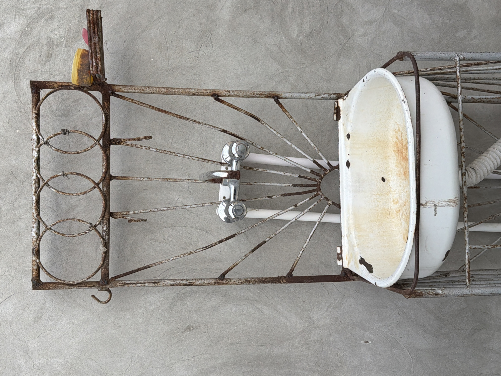 A rusty metal frame, likely part of an outdoor sink stand, is positioned against a textured, light gray concrete wall. The frame is designed with circular decorative elements at the top and radiating spokes that support a white, chipped enamel basin. A chrome faucet with two handles is mounted above the basin. On a small shelf attached to the right side of the frame, a yellow bar of soap and a pink object, possibly another bar of soap or a sponge, are visible. The overall impression is one of disuse and weathering, suggesting it is an outdoor fixture in a state of disrepair. This scene is located in the city of Sabirabad, Azerbaijan.