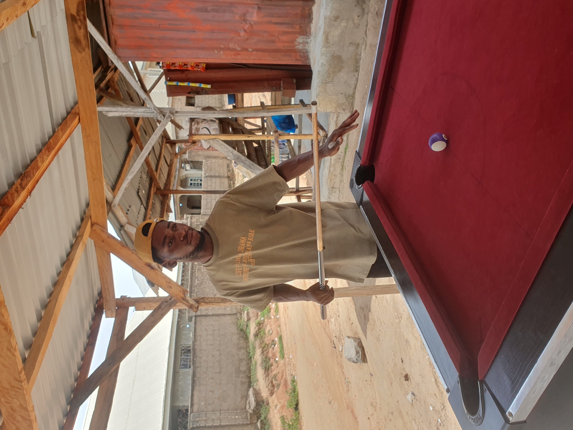 A young Black man wearing a tan t-shirt and a yellow baseball cap stands over a red pool table outdoors in Lokoja, Nigeria. He holds a pool cue with his right hand, preparing to strike a purple and white striped pool ball. The pool table is positioned on a dirt surface under a partially constructed wooden and metal roof. In the background, there are buildings, including a house with grey brick walls and a corrugated metal structure. The scene appears to be an open-air recreational area.
