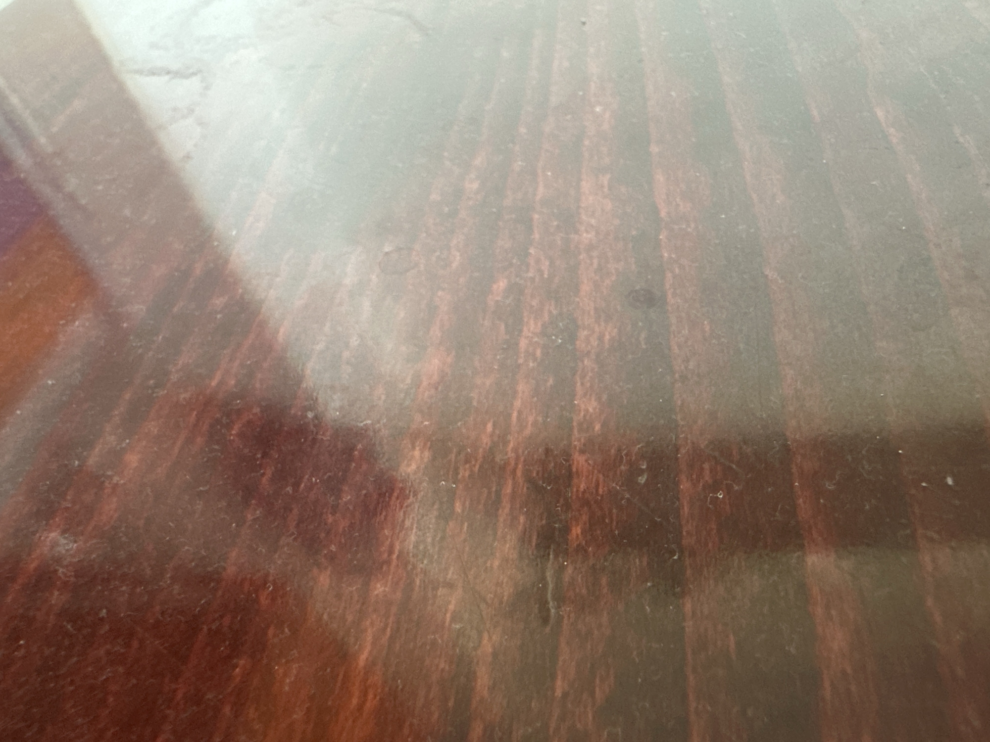 The image displays a close-up, angled view of a polished wooden surface, likely a table or desk. The wood has a deep reddish-brown color with visible grain lines running vertically. The surface exhibits a high gloss, reflecting light and creating areas of bright glare, particularly in the upper left portion of the image. There are faint smudges and dust particles scattered across the surface, indicating it is not perfectly clean. The overall impression is of a smooth, hard, and slightly aged wooden material. The location in Zvezdara Urban Municipality, Serbia, is not discernible from the visual information provided in the image.