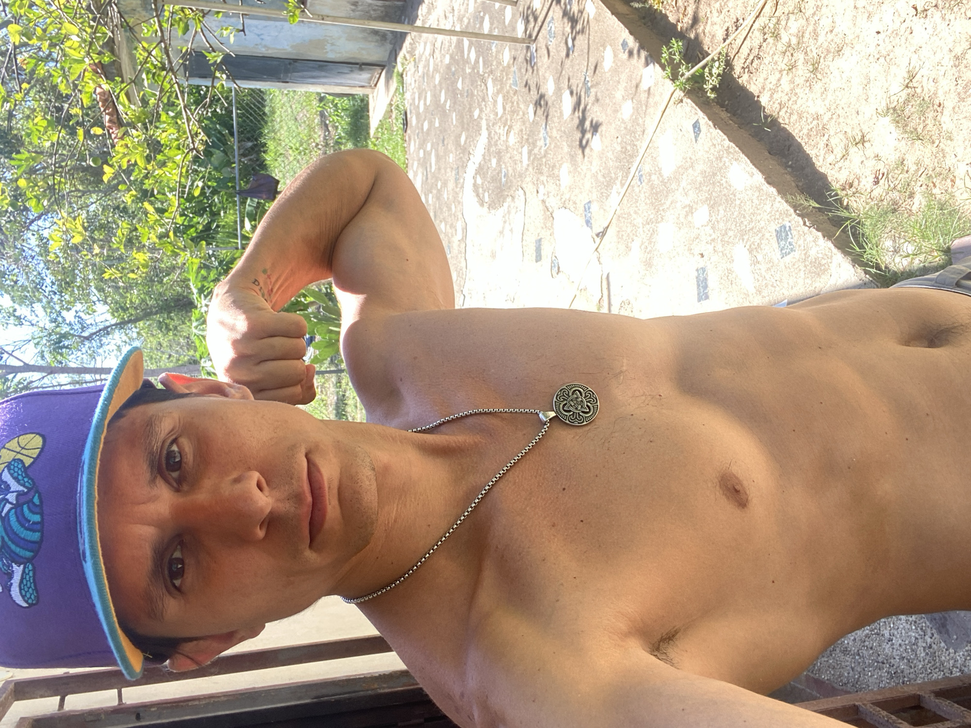 A shirtless man wearing a purple baseball cap with a yellow bill is shown in a medium shot, flexing his left arm. He is wearing a silver necklace with a circular pendant. The location appears to be outdoors, with a concrete surface visible and foliage in the background. The setting is indeterminate in Los Velásquez, Venezuela.