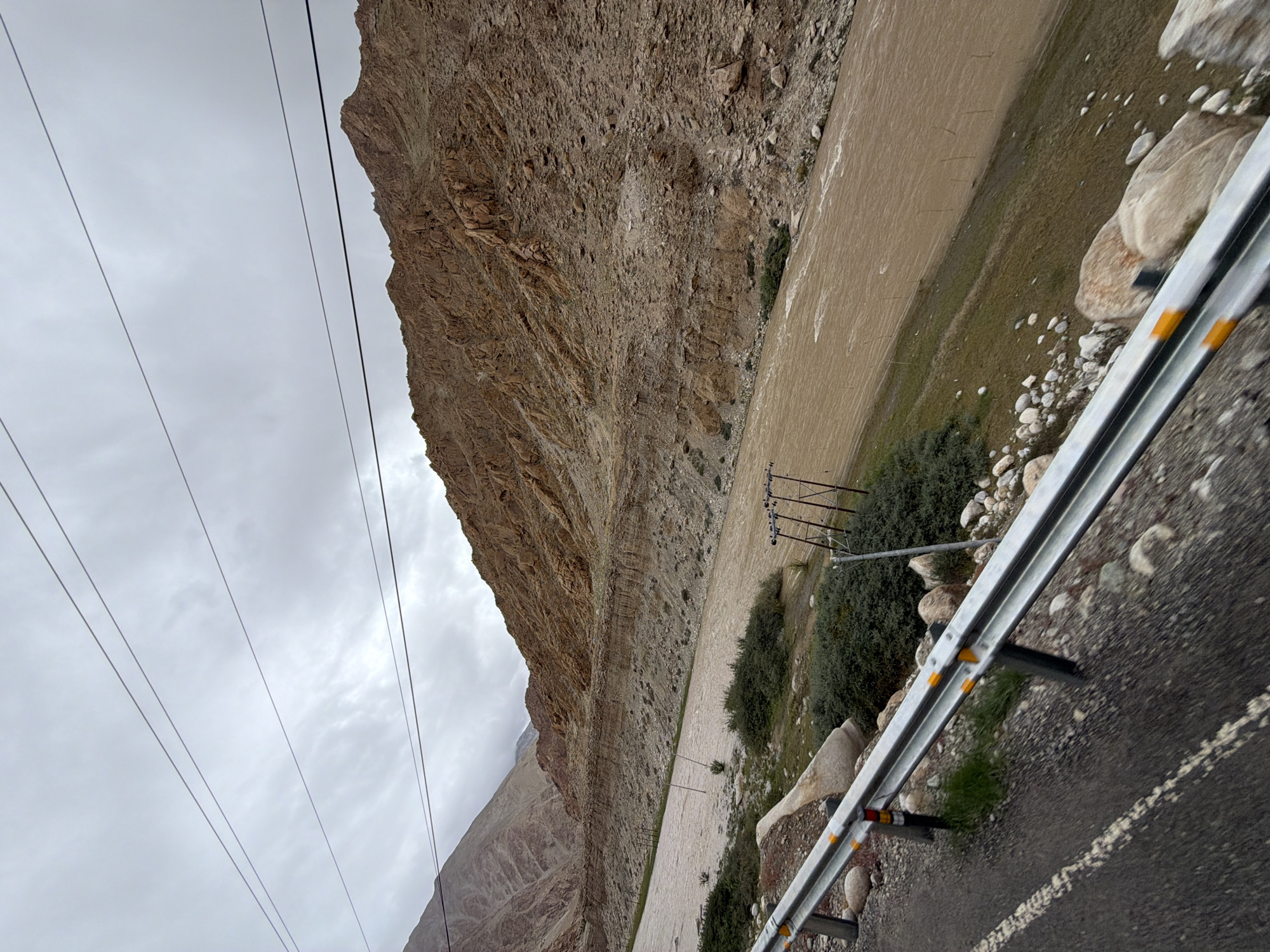 The image depicts a wide, muddy river flowing through a valley with steep, barren, rocky hills on either side. The sky is overcast with gray clouds. In the foreground, a road runs along the left side of the frame, bordered by a metal guardrail with yellow reflectors. The road surface is asphalt with a white dashed line. The river occupies the middle ground, with patches of green vegetation and scattered rocks along its banks. In the background, the rugged, brown mountains dominate the landscape. Several utility poles with wires are visible near the riverbank. The scene appears to be in a remote, mountainous region. The location is identified as Gayk, India.