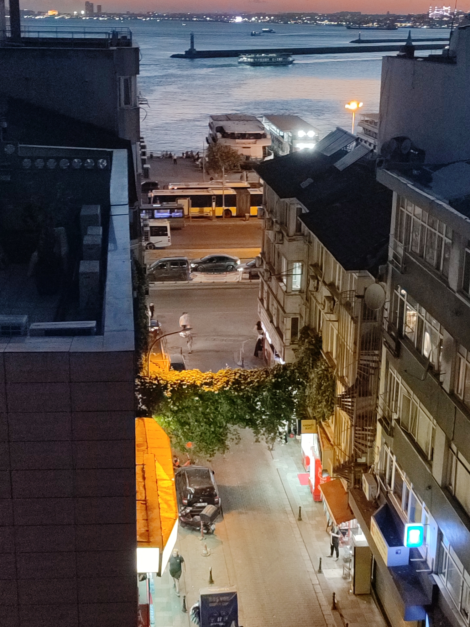 This is a medium-angle, high-quality, vertical, full-color image taken at dusk from an elevated perspective, looking down onto a street in Kadıköy, Turkey. The scene captures a bustling urban environment with elements of both a waterfront and a street scene.
In the background, a body of water, likely the Bosphorus Strait, is visible. Several boats, including a ferry and a lighthouse, are present on the water. Distant city lights and structures are visible on the opposite shore.
The foreground and midground focus on buildings lining a street. The buildings are multi-story structures with visible windows, balconies, and architectural details such as spiral staircases and air conditioning units. Some buildings have businesses on the ground floor with illuminated signs and displays.
The street is paved and appears to be well-lit by streetlights and the lights from the buildings. Several vehicles, including cars and buses, are parked or driving on the street. Pedestrians are also visible walking on the sidewalks and in the street.
The overall impression is of a vibrant city scene at twilight, showcasing the blend of urban development, transportation, and everyday life in Kadıköy. The lighting suggests the transition from day to night, with warm tones from artificial lights contrasting with the cooler blues and purples of the sky and water.