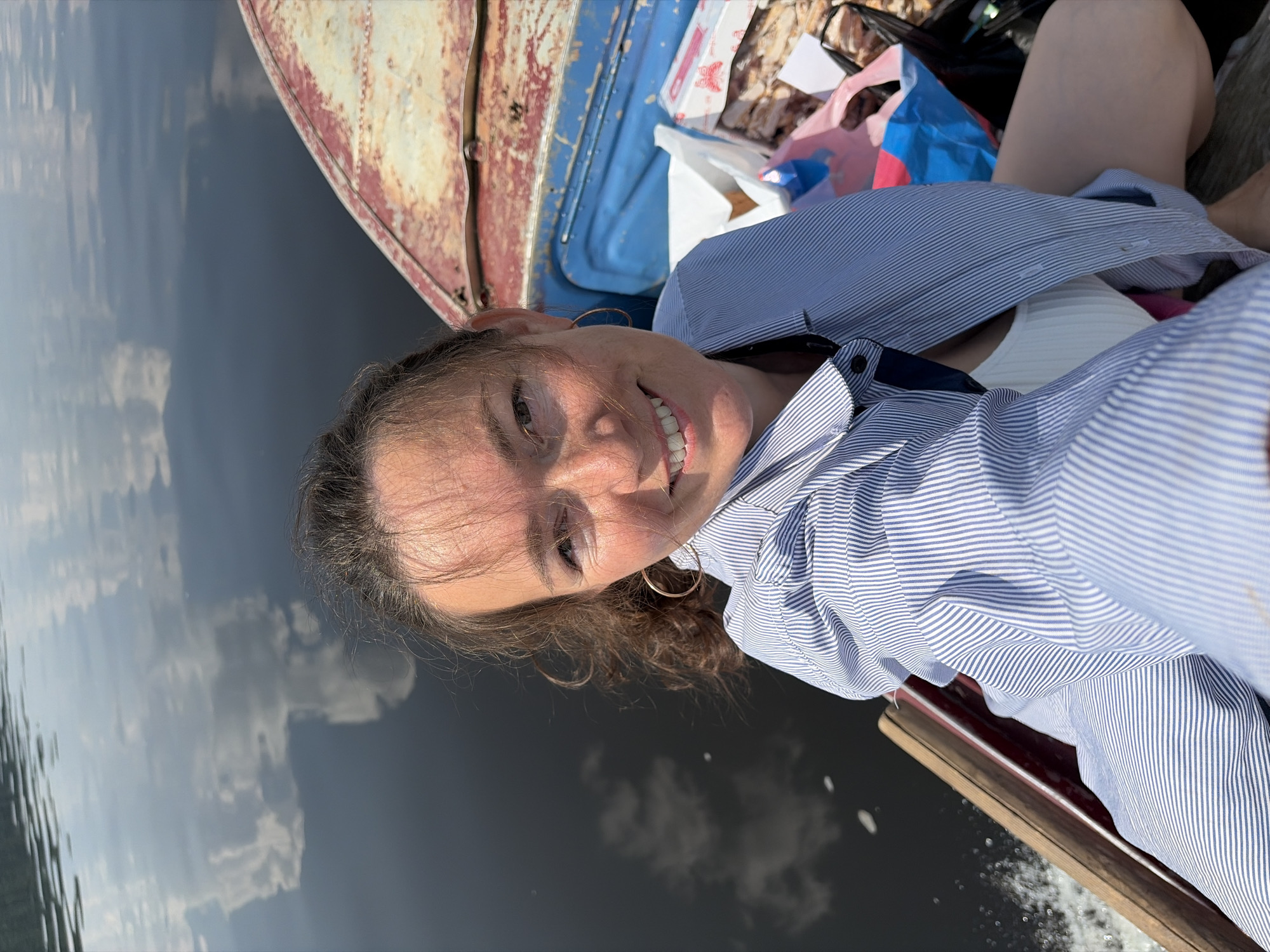 A woman with brown hair and hoop earrings, wearing a blue and white striped button-down shirt over a white ribbed tank top, is taking a selfie while sitting in a boat on a body of water. Her reflection and the reflection of clouds are visible on the dark water. The boat appears to be made of wood and has a weathered, rusty red and blue interior. Several bags and boxes of food are visible in the boat behind her. The environment suggests an outdoor recreational activity on a lake or river. The image is taken from a high angle, looking down at the woman.