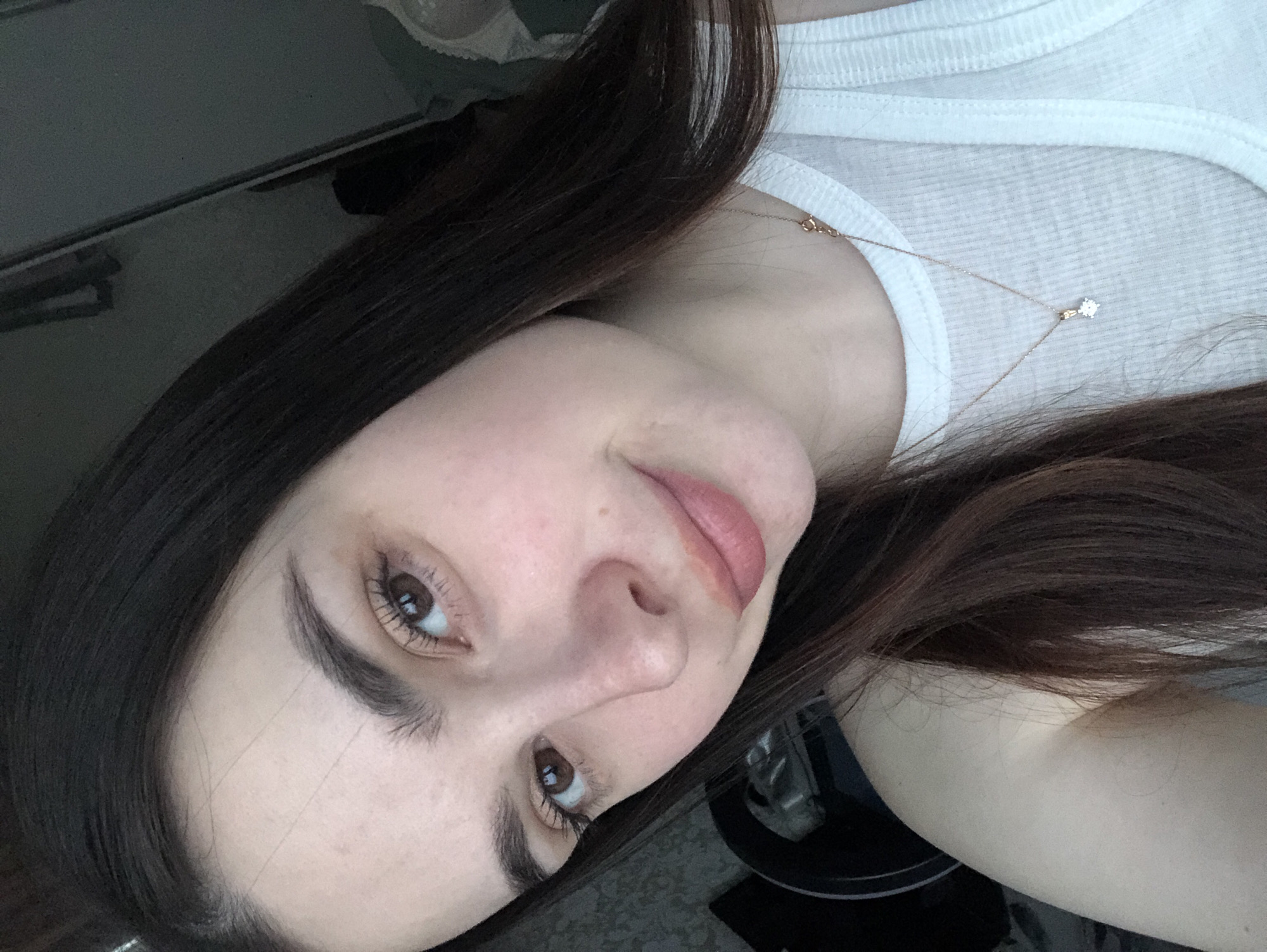 A selfie of a woman with long, dark brown hair. She is wearing a white tank top and a delicate gold necklace with a small pendant. The background is blurry and indistinct, but includes a dark, possibly metallic surface. There is no information in the image to connect it to the city of Noviy, Russia.