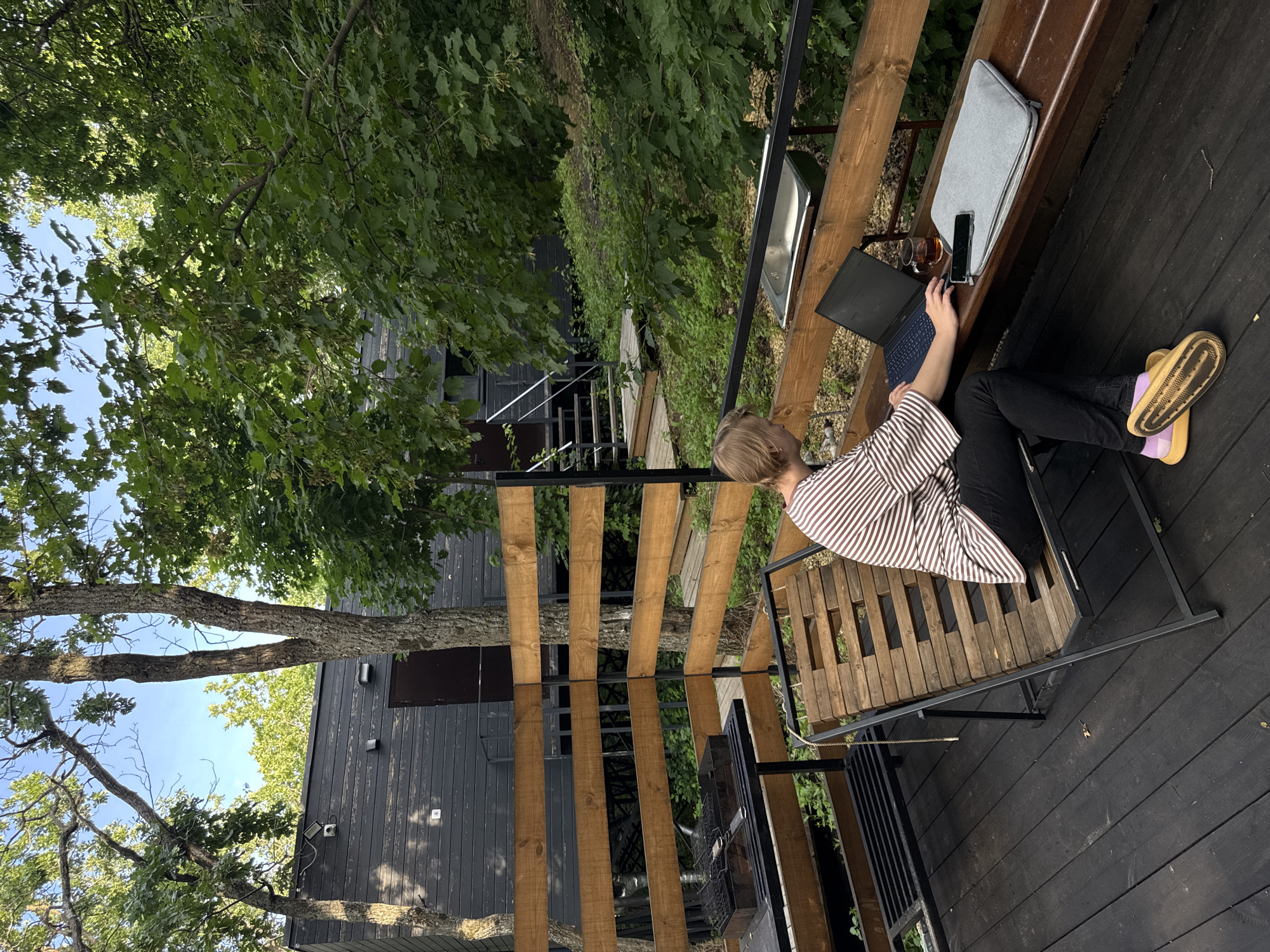 A person with blond hair, wearing a striped shirt and dark pants, sits on a slatted wooden lounge chair on a dark wooden deck. They are working on a laptop, with a glass of tea and a smartphone on a grey laptop sleeve next to them on a wooden bench. The deck overlooks a natural setting with trees and foliage. In the background, a dark, modern building with wooden accents and stairs leading up to it is visible, situated within a densely wooded area. There is also a barbecue grill on the deck. The location is identified as Ust-Kurdyum, Russia.