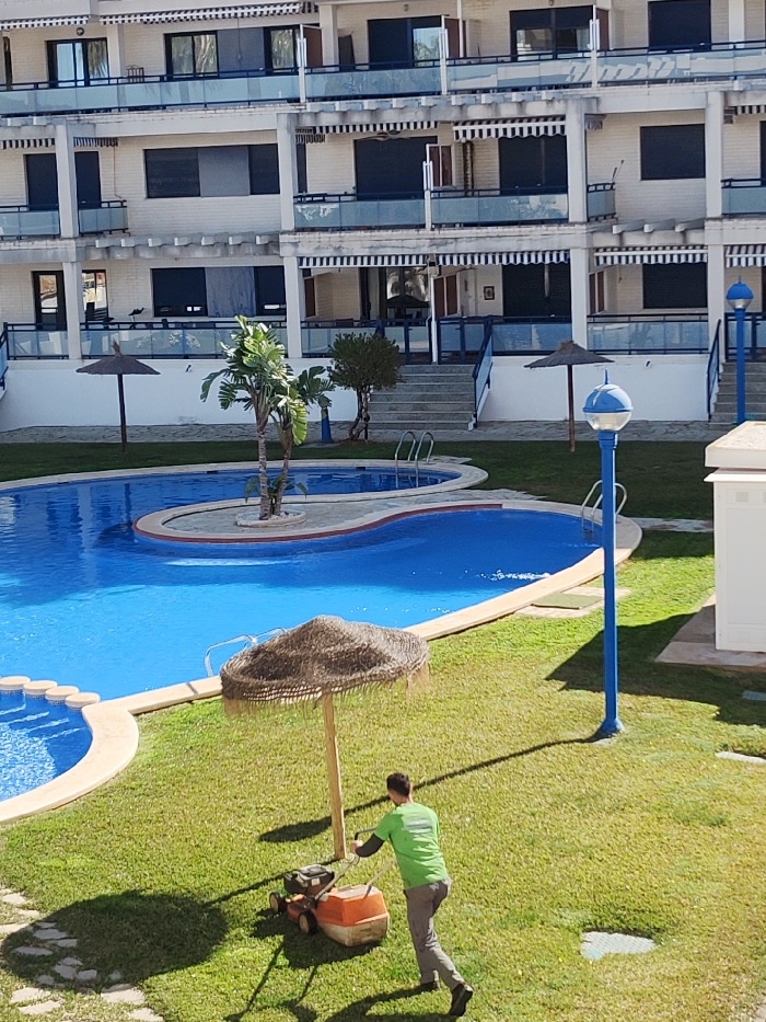 The scene depicts an outdoor common area of a residential or resort complex in Rotes, Spain. A multi-story building forms the backdrop, characterized by white facades, multiple balconies with blue railings, dark-shuttered windows, and horizontally striped awnings. In the midground, a large, irregularly shaped swimming pool with bright blue water is visible, featuring light-colored coping and steps. A small island within the pool hosts a palm tree. The foreground consists of a well-maintained green lawn where a male individual is actively pushing an orange and black gasoline-powered lawn mower. The man wears a bright green t-shirt and grey pants and is moving rightward across the grass. Several thatched umbrellas are positioned around the pool and lawn. Blue-poled street lamps are also present. Distinct shadows on the grass and structures indicate bright sunlight. Steps and walkways lead from the ground level to various entrances of the building.