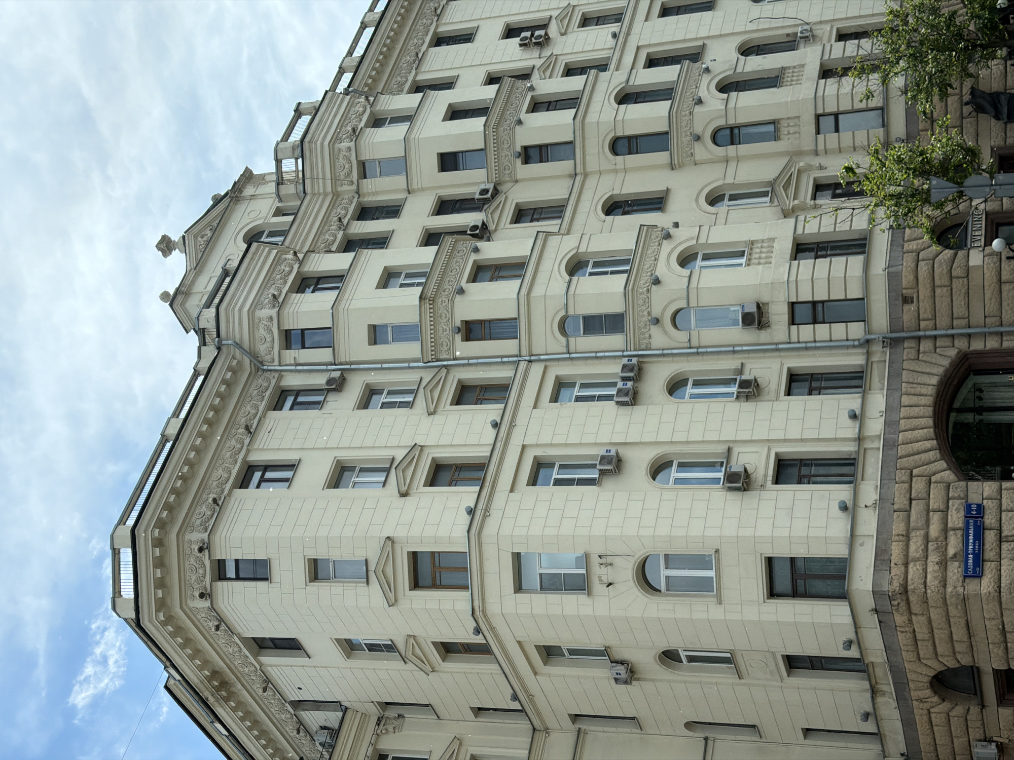 An upright view captures the ornate facade of a large building in Moscow, Russia, under a partly cloudy sky. The multi-story structure is predominantly cream-colored, featuring a classical or Stalinist Empire architectural style with rich decorative elements.
The main section of the building displays a complex arrangement of windows, including numerous rectangular, arched, and semicircular designs. Many windows are fitted with external air conditioning units. The facade is adorned with intricate moldings, cornices, and decorative panels, giving it a grand and detailed appearance. Vertical sections between windows feature patterned reliefs. Balconies with metal railings are visible on the upper levels.
On the right side, the building transitions to a section clad in rough-hewn stone blocks, featuring large arched openings. Green foliage from trees partially obscures this section.
Visible text on a blue street sign attached to the stone facade reads "САДОВАЯ-ТРИУМФАЛЬНАЯ" (Sadovaya-Triumfalnaya), "4-10", and "ТВЕРСКАЯ" (Tverskaya). Another sign, partially visible and inverted in the original image, appears to read "EVENING" on the black background of the stone section.
The image depicts a daytime scene, with natural light illuminating the building's architectural details. No people or activities are visible, focusing solely on the urban architecture.
