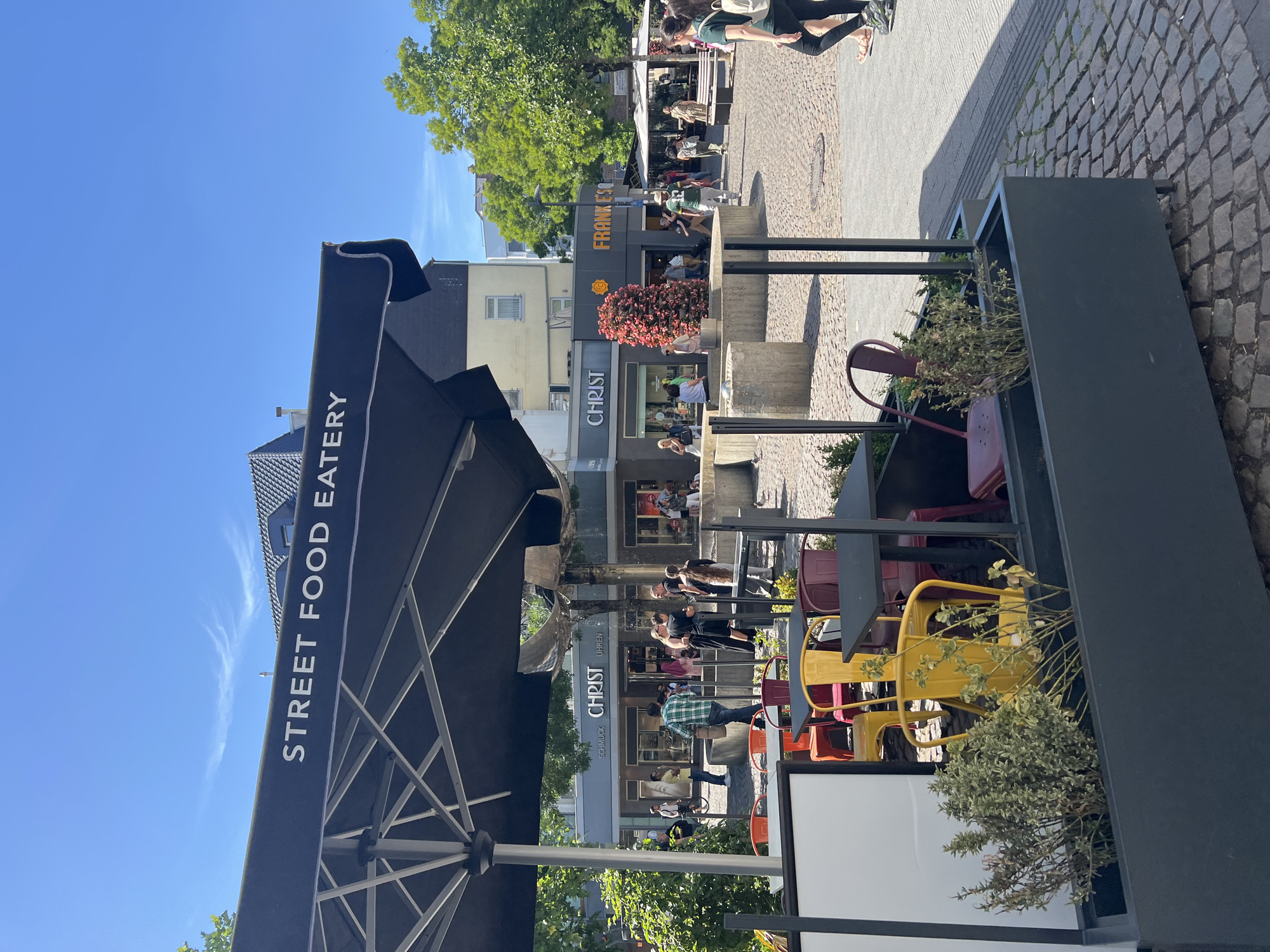 The image captures a sunlit street scene in Aachen, Germany, likely during midday given the bright sunlight and clear blue sky with a few wispy clouds. The perspective is from a slightly elevated viewpoint, looking down onto an outdoor seating area and the street beyond.
In the foreground, a large black umbrella with the white text "STREET FOOD EATERY" emblazoned on its side dominates the left side of the frame. Adjacent to it is a dark grey planter filled with greenery. Behind this, a row of shops lines the street. One shop clearly visible has a sign reading "FRANKFURTER" with a stylized orange logo. Another shop's sign displays "CHRIST SCHMUCK UHREN," indicating it sells jewelry and watches.
The street is paved with cobblestones. Several people are visible, both sitting at outdoor tables and walking along the street. Some are patrons of the eateries, while others appear to be shoppers or passersby. The people are casually dressed, suggesting a relaxed, everyday atmosphere. The overall mood is bright and cheerful, amplified by the sunny weather. Several colorful metal chairs are scattered around the outdoor seating areas, adding a touch of vibrancy to the scene. The presence of trees with lush green foliage suggests a warm season, likely late spring or summer.