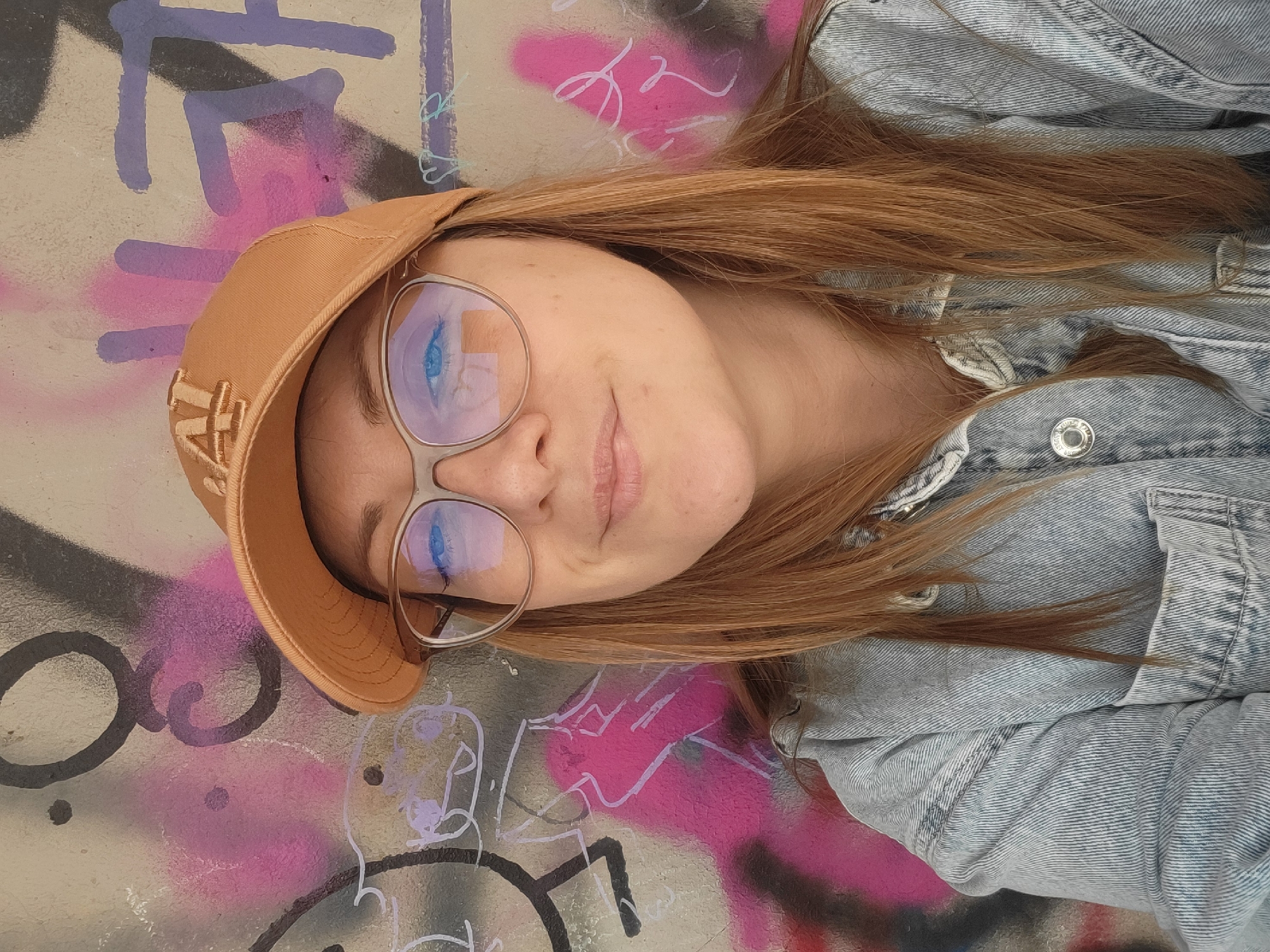 A young woman wearing a tan baseball cap with "NY" embroidered on the front, and wire-rimmed glasses, lies on a graffiti-covered surface. She is wearing a light-blue denim jacket. The graffiti includes black, purple, pink, and white paint. The scene appears to be in an urban environment, likely in Budva, Montenegro, but specific location details cannot be determined from the image.