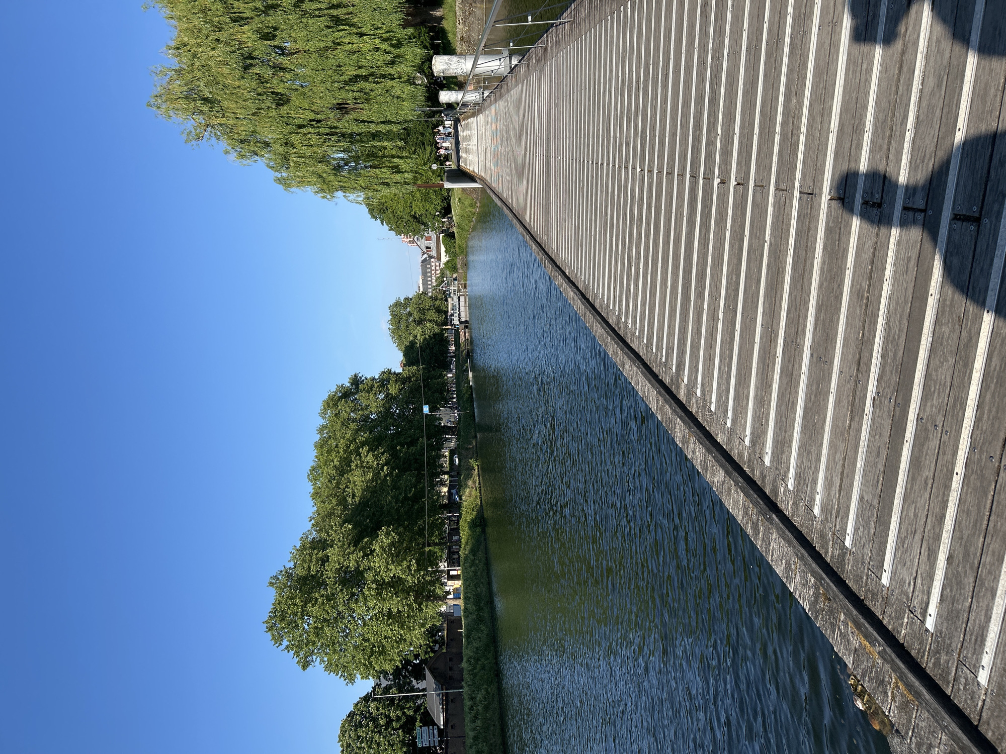 The image shows a wooden bridge over a canal in Strasbourg, France. The bridge is made of planks with white stripes. The canal is narrow, and there are trees and buildings along the banks. The sky is blue. A person's shadow is visible on the bridge.
