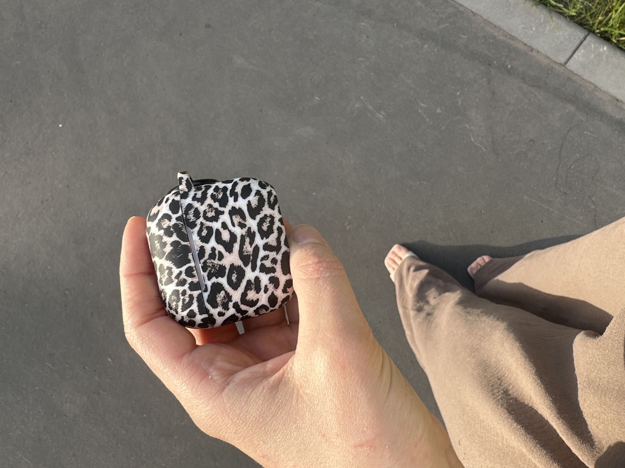 The image shows a person holding a leopard-print AirPods case in their hand. The person is outdoors, standing on an asphalt surface, possibly a sidewalk or path. They are wearing light brown trousers. Their bare feet are visible, suggesting warm weather. In the background, a strip of green grass and a concrete curb indicate an urban or park-like setting. The sunlight casts shadows, indicating it is likely daytime. The scene appears casual and relaxed. There is no visible text in the image.