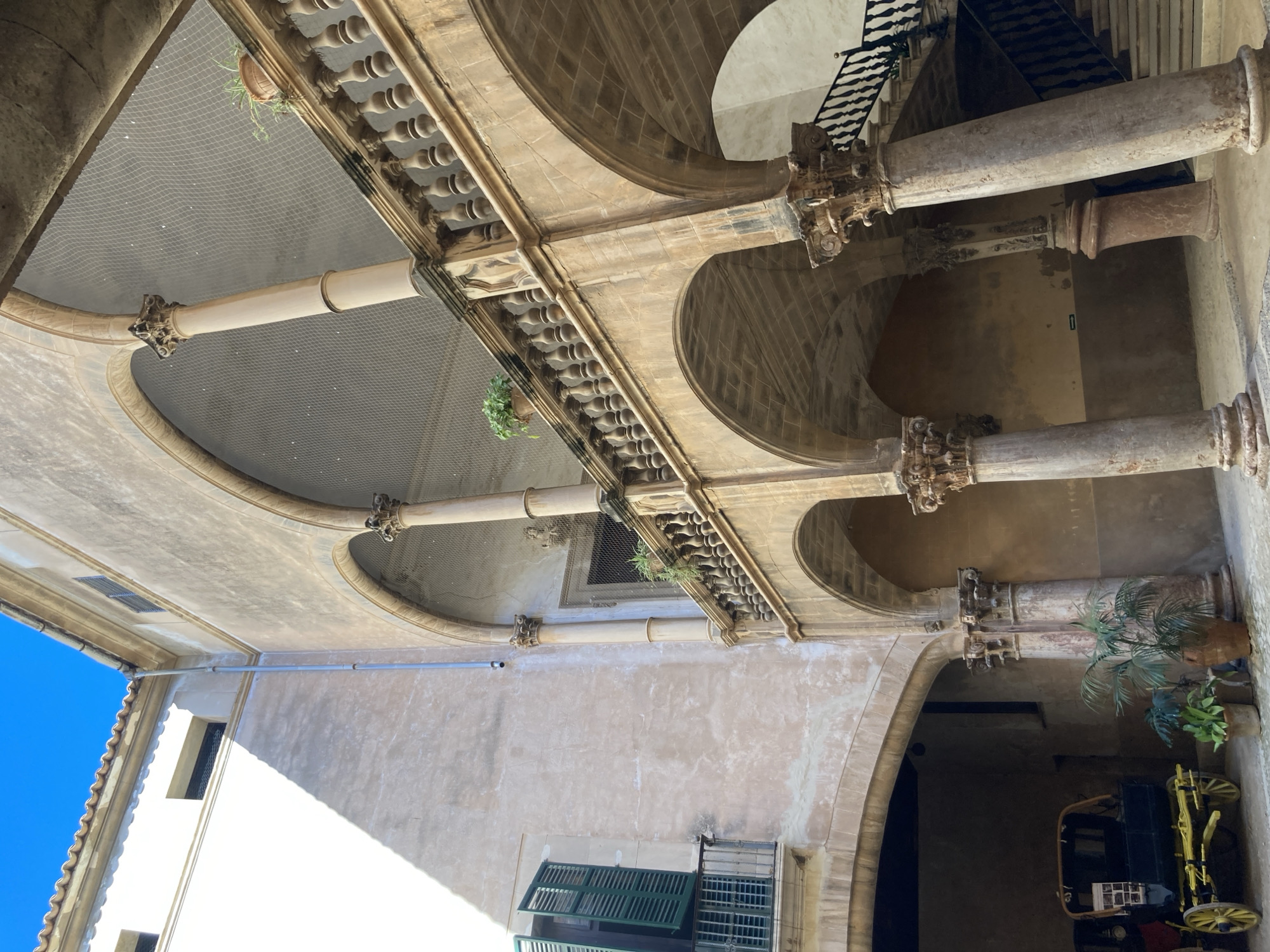 This is a detailed, matter-of-fact description of a courtyard in Palma de Mallorca, Spain, specifically within the courtyard of the historic Palau March.
The scene depicts a sunlit courtyard with arched walkways and a prominent staircase. The architecture features classical elements, including large stone columns with ornate capitals and stone balustrades. The walls are plastered and weathered, showing signs of age. Above the arches, a fine mesh netting is visible, likely to prevent birds from roosting.
In the lower left portion of the courtyard, a vintage carriage is parked. It is dark-colored with yellow wheels and appears to be an exhibit or decorative element. Several potted plants, including palm fronds, are placed near the carriage and along the base of the columns.
To the left, a building facade is visible with a window featuring green shutters that are partially open. A small balcony with a metal railing extends from this window. The sunlight casts strong shadows across the facade, highlighting the textures and architectural features.
The staircase is made of stone with dark metal railings and leads upwards along the right side of the courtyard, disappearing into the upper levels. The overall impression is one of historical grandeur and a peaceful, sheltered outdoor space within a larger building complex. The sky above is a clear, bright blue.