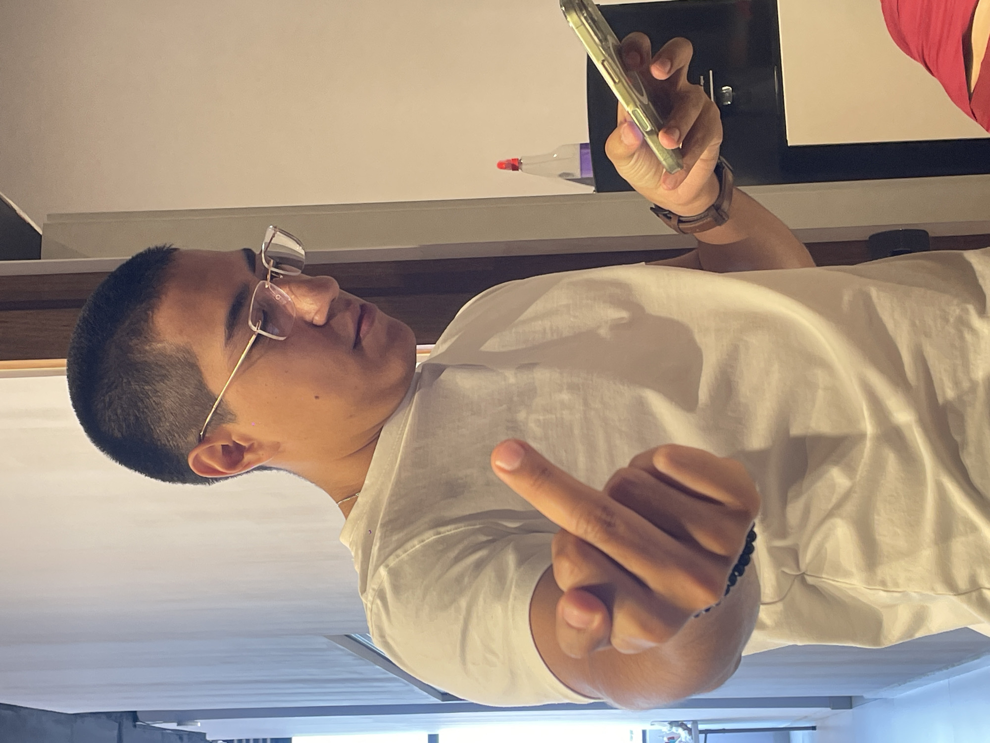 A medium shot presents a male individual, identifiable by his closely cropped dark hair and light complexion. He is wearing eyeglasses and a white t-shirt. The man is holding a smartphone in his right hand, while his left hand is raised, displaying the middle finger. The photograph is indoors, with a section of a television or monitor visible in the background. The location appears to be in Mexico City, Mexico.